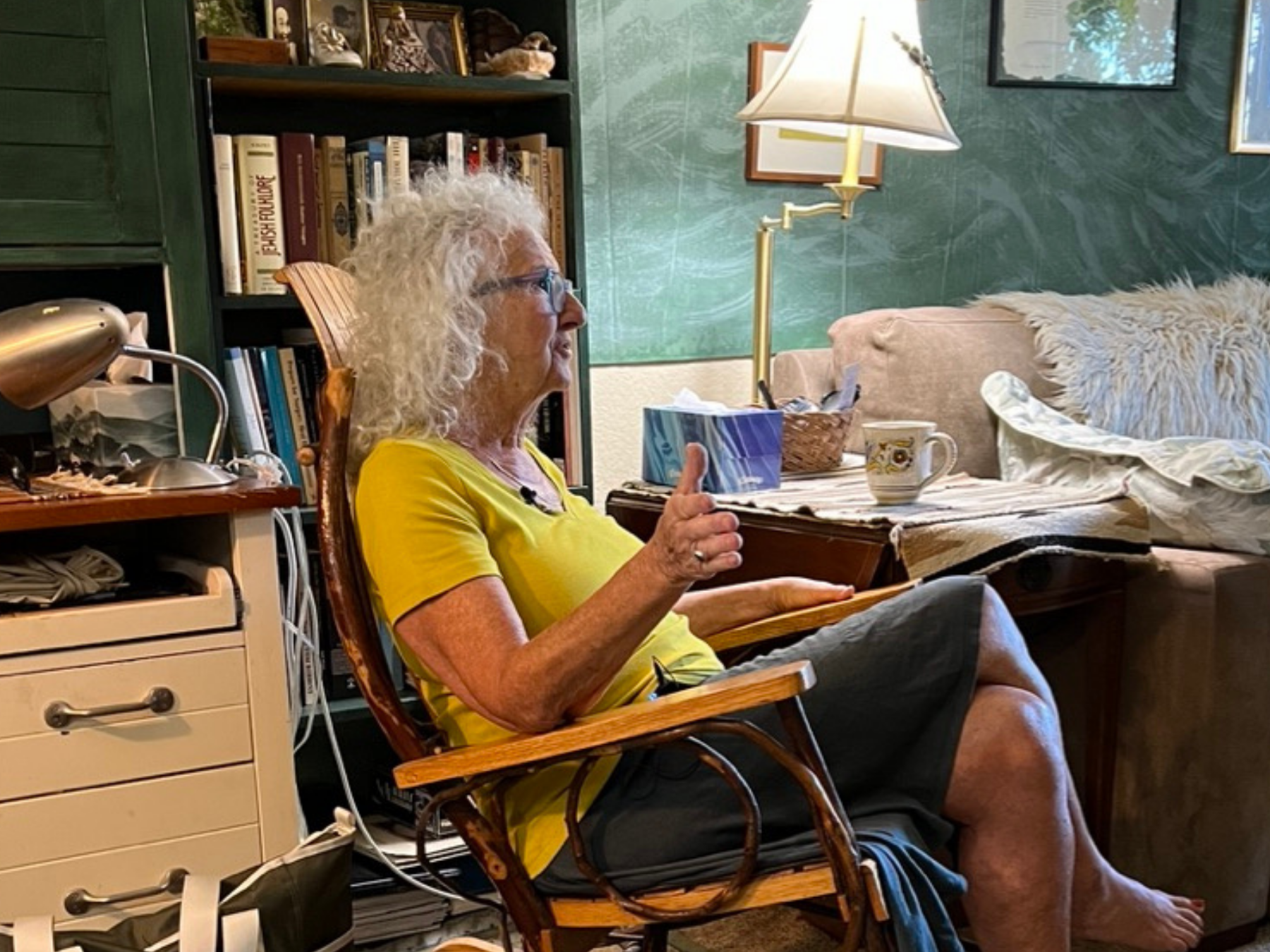 An elderly woman with curly white hair and glasses sitting on a wooden rocking chair in a cozy living room, gesturing with her hand while speaking.