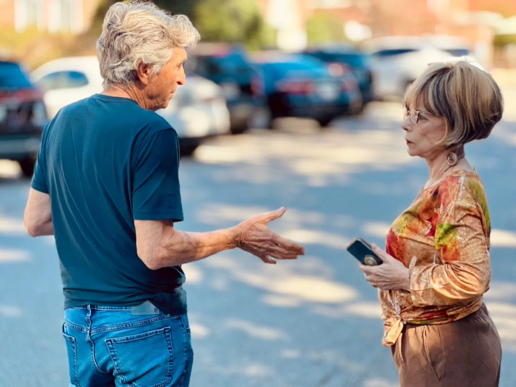 An elderly man and woman are engaged in a conversation in a parking lot. The man has gray curly hair, is wearing a blue shirt, and gesturing with his hands. The woman has short gray hair, is wearing glasses, a multicolored top, and holding a smartphone. Cars are parked in the background.