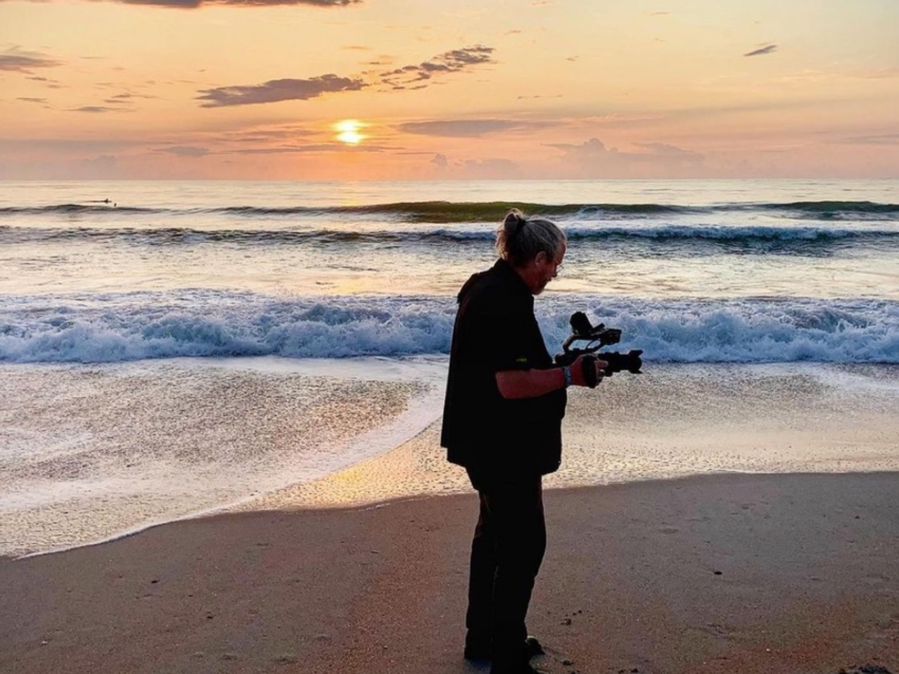 A person with gray hair tied in a bun, dressed in black, is standing on the beach holding a camera or filming device, with waves and a sunset in the background.
