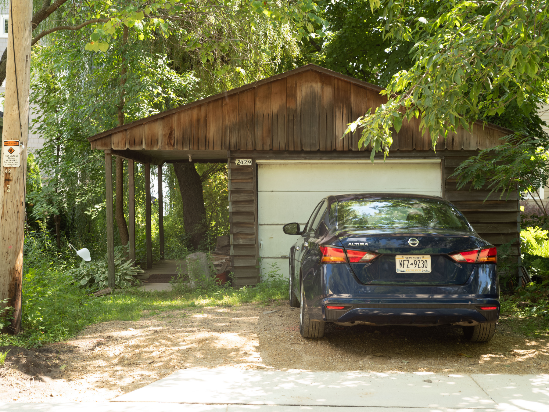 A black Nissan Altima parked in front of a small, rustic wooden garage with a white roll-up door, surrounded by green trees and plants, with a gravel driveway and a wooden utility pole on the left.
