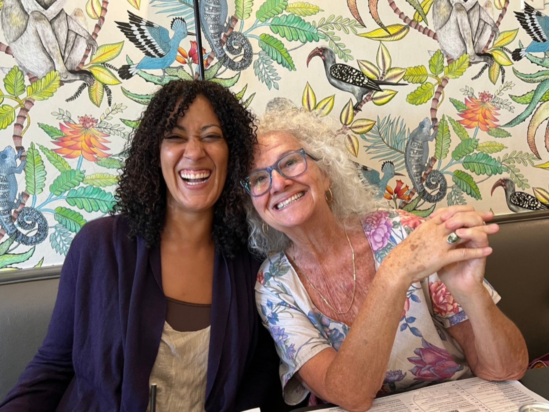 Two women smiling and laughing at a table with a colorful tropical bird and plant wallpaper in the background.