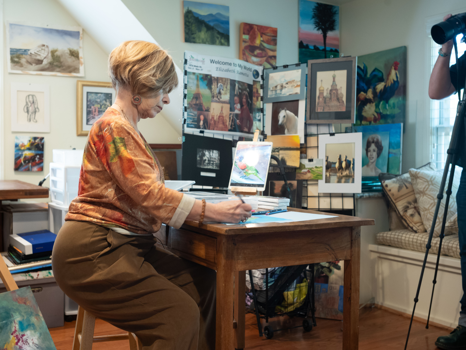 An older woman with short light hair, wearing a patterned blouse and brown pants, sitting on a stool at a wooden table in an art studio. She is working on her art surrounded by paintings and art supplies, with a camera person recording her.