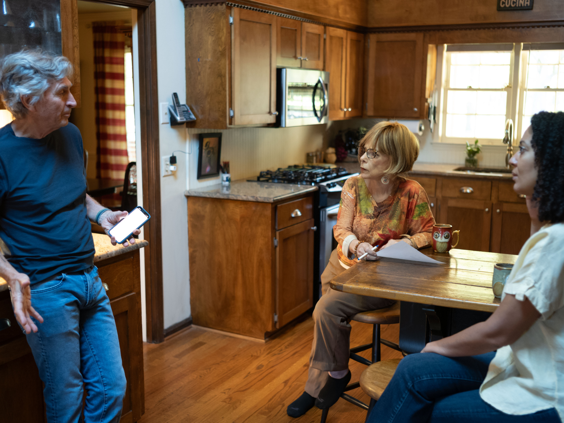 Three women having a conversation in a kitchen, one of them standing holding a phone, two seated at a table with mugs, wooden cabinets, a stove, and a window in the background.