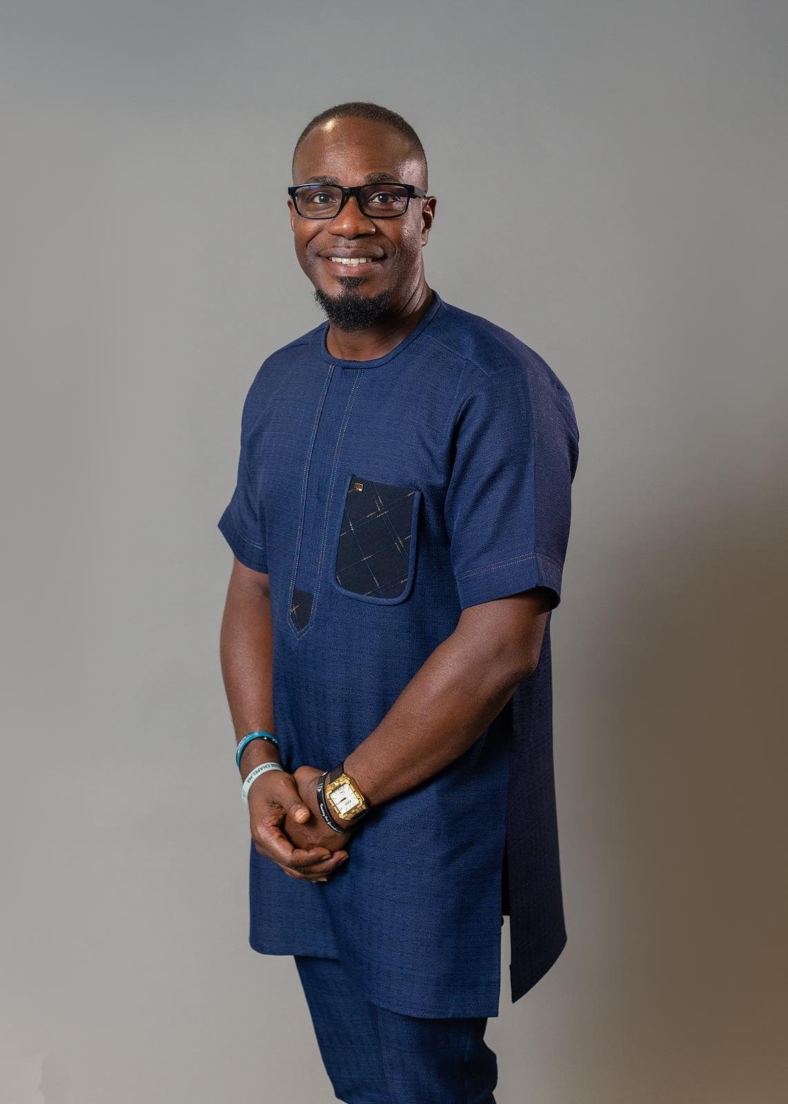 Portrait of our Senior Pastor smiling with glasses, wearing a blue traditional African outfit, standing with hands clasped against a neutral background.