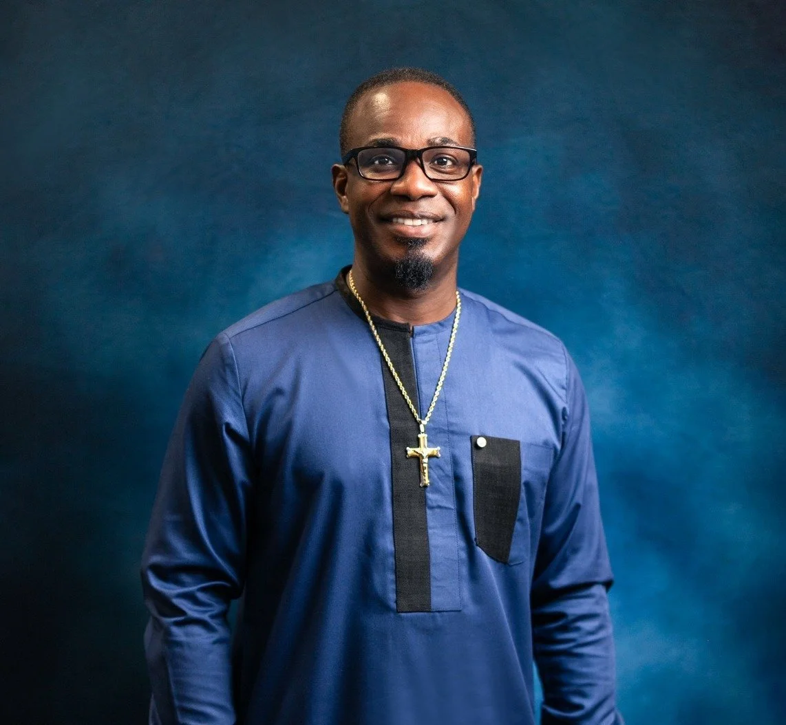 A man wearing glasses and a blue traditional African outfit with black accents, a gold chain with a cross, standing against a dark blue background.