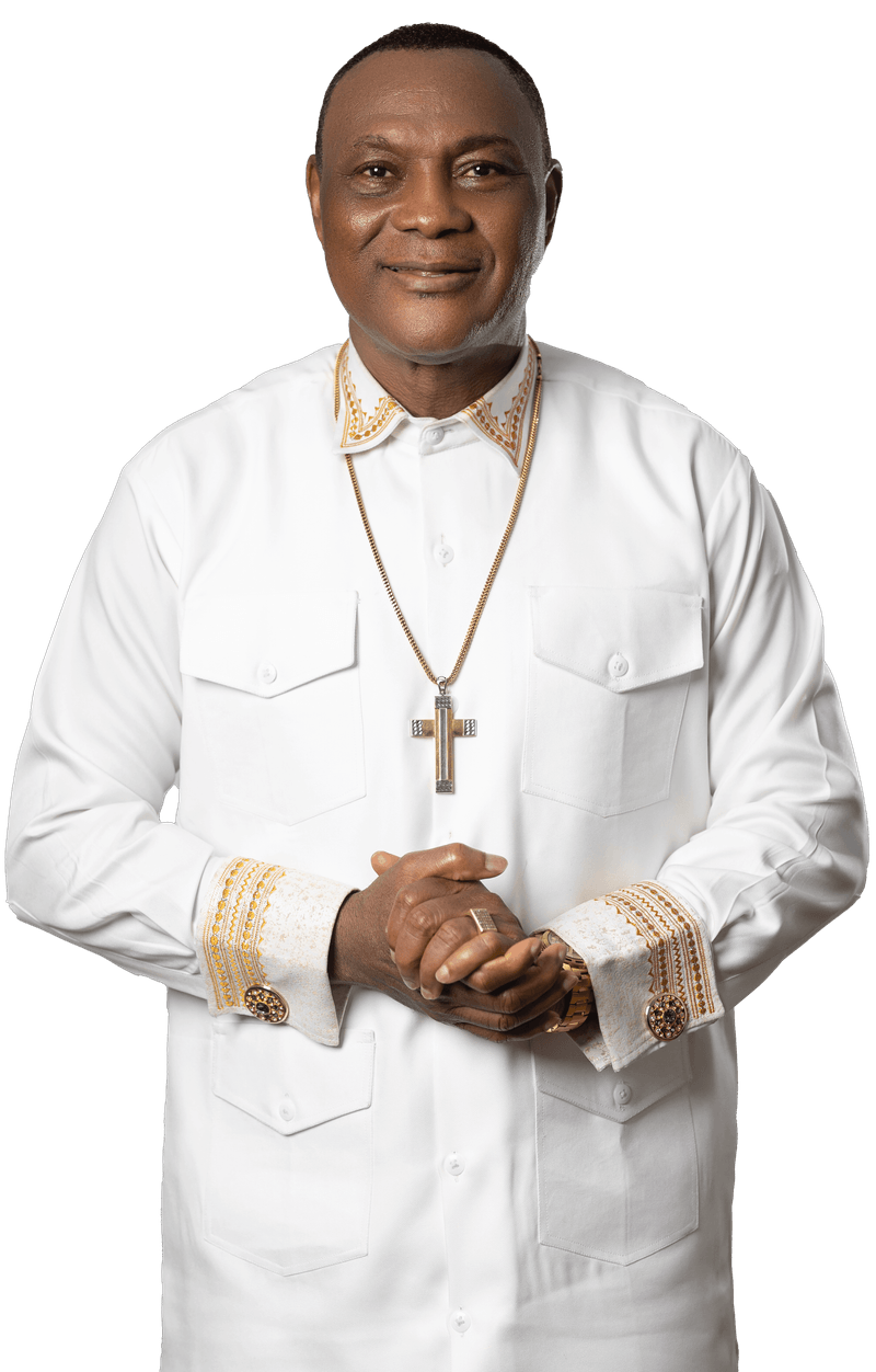 A man wearing a white shirt with gold embroidered details on the collar and cuffs, and a gold cross necklace. He is smiling with his hands clasped in front of him.