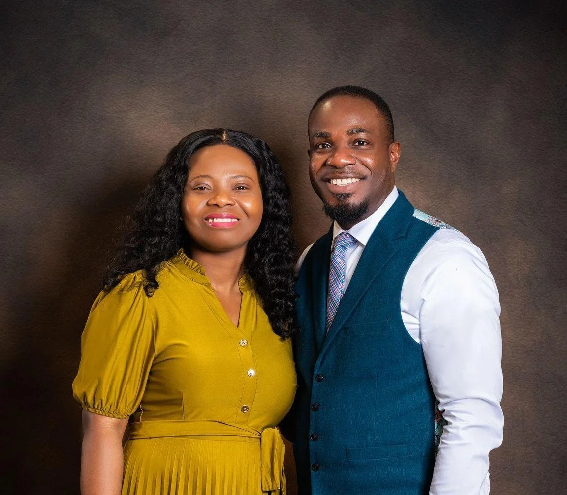 A smiling couple standing closely together, the woman wearing a mustard yellow dress and the man in a white shirt with a teal vest and tie, against a plain brown background.