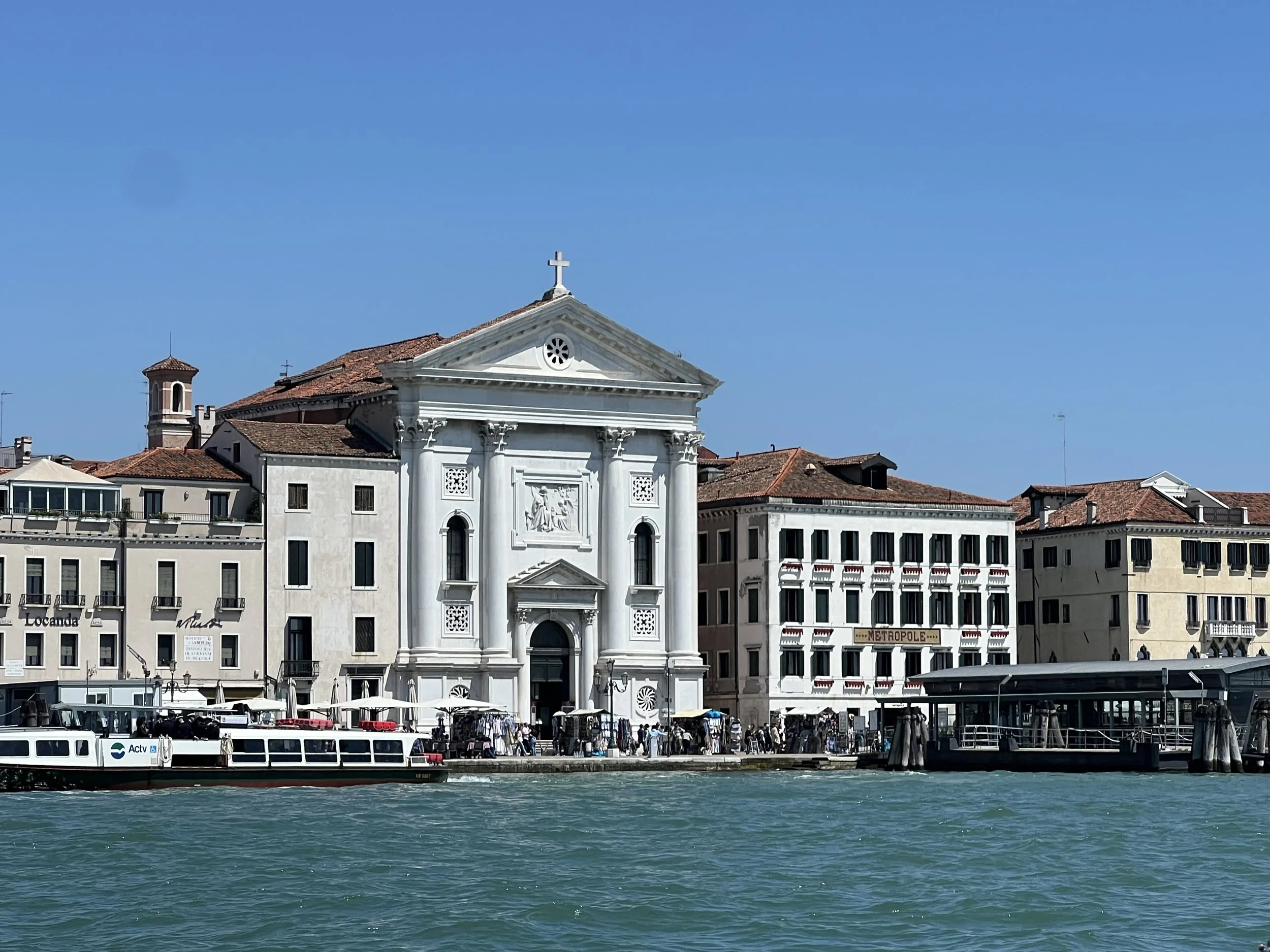 View of a historic church with a cross on top, situated along a waterfront with boats and apartment buildings in Venice, Italy.