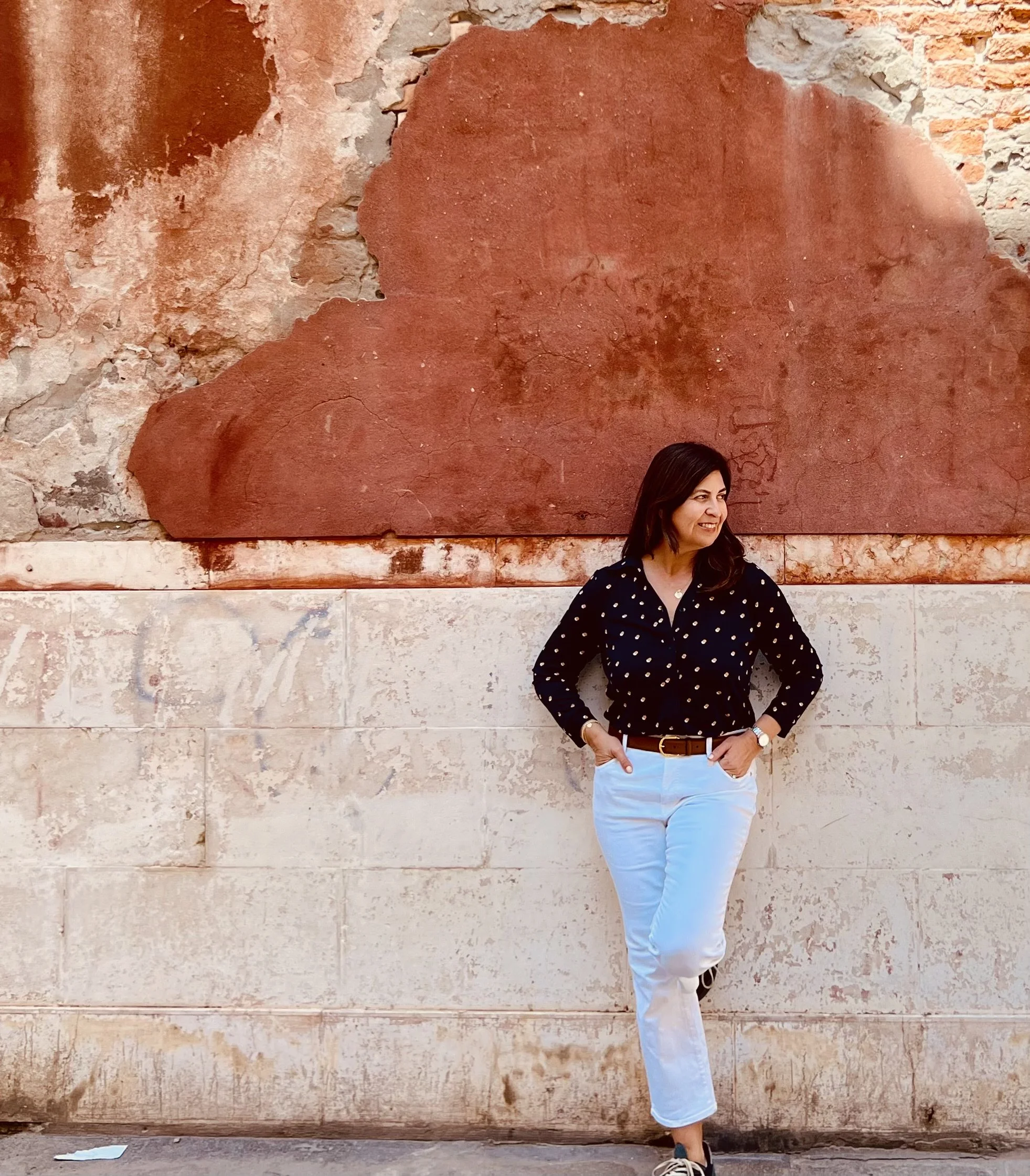 A woman standing against a weathered brick wall with a large abstract red mural. She is wearing a black polka dot shirt, light blue pants, and has her hands in her pockets, smiling and looking to her left.