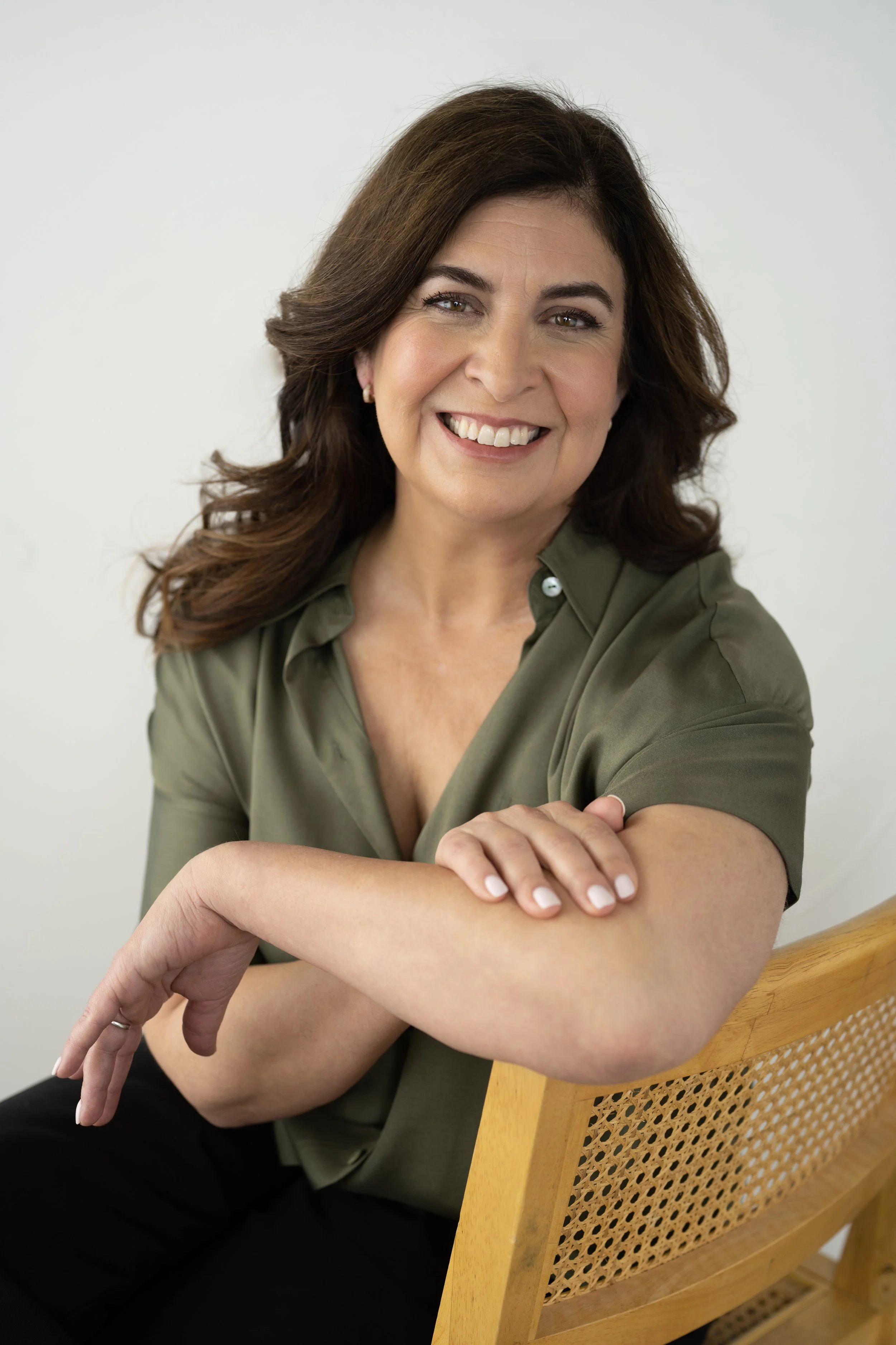 A smiling woman with dark brown hair, wearing an olive green short-sleeve shirt, leaning on a wooden chair with a woven back, against a plain light background.