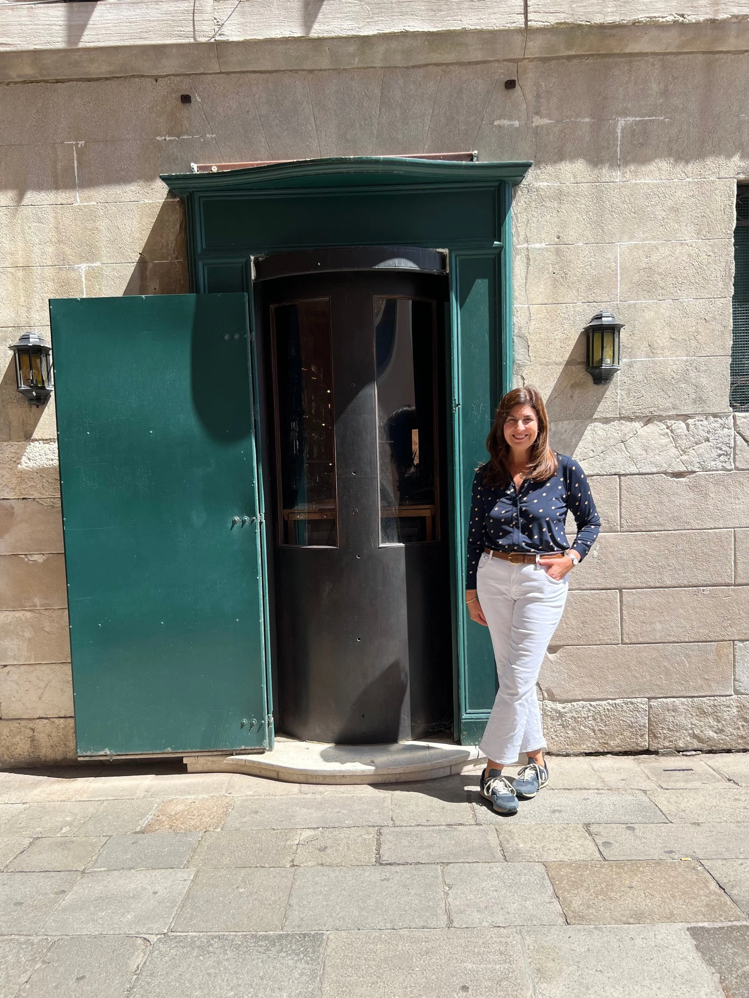 A woman with brown hair wearing a navy blue polka dot blouse, white pants, and sneakers standing outside a building with a stone facade, smiling while posing next to a small green door with open shutters.