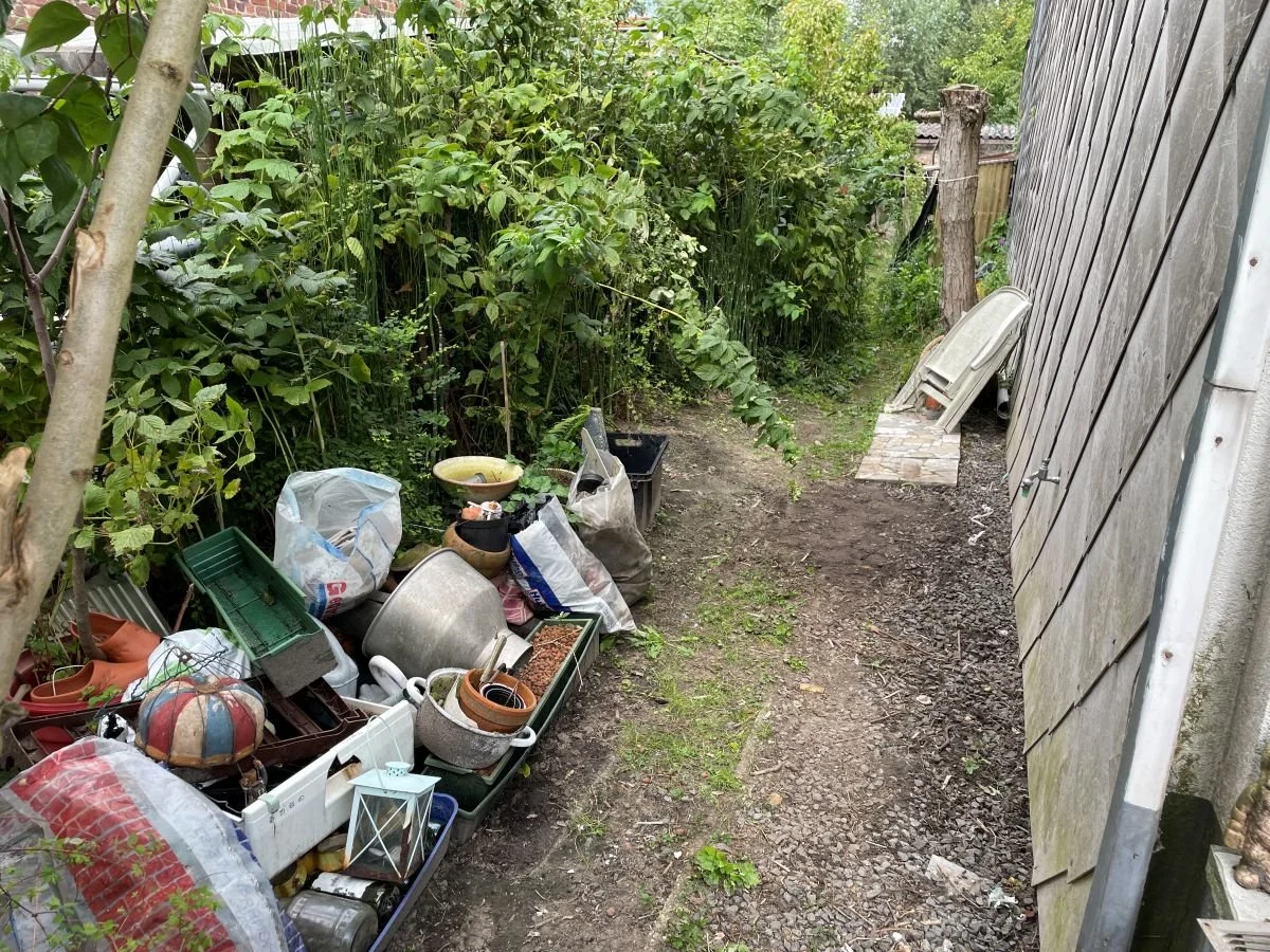 Een tuinpad met tuinplanten en een hoop afval en tuingereedschap aan de zijkant naast een schutting.