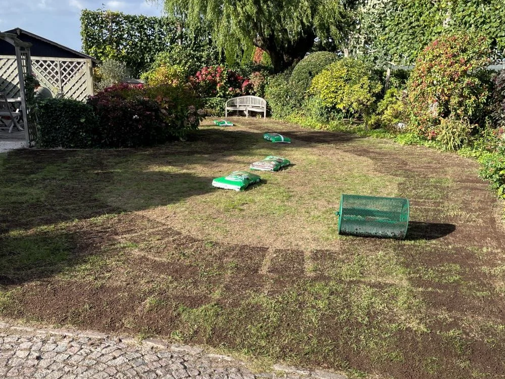 Tuin met liggende tuinmaterialen zoals aardezakken, een compostroller en een bankje, omgeven door bloemen en struiken.
