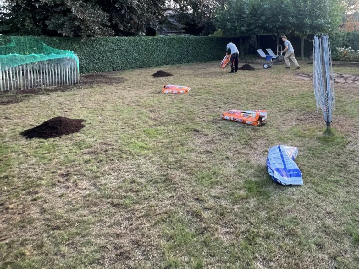 Twee mensen werken in een tuin met nieuwe graszoden en gereedschap, omringd door bomen en struiken.