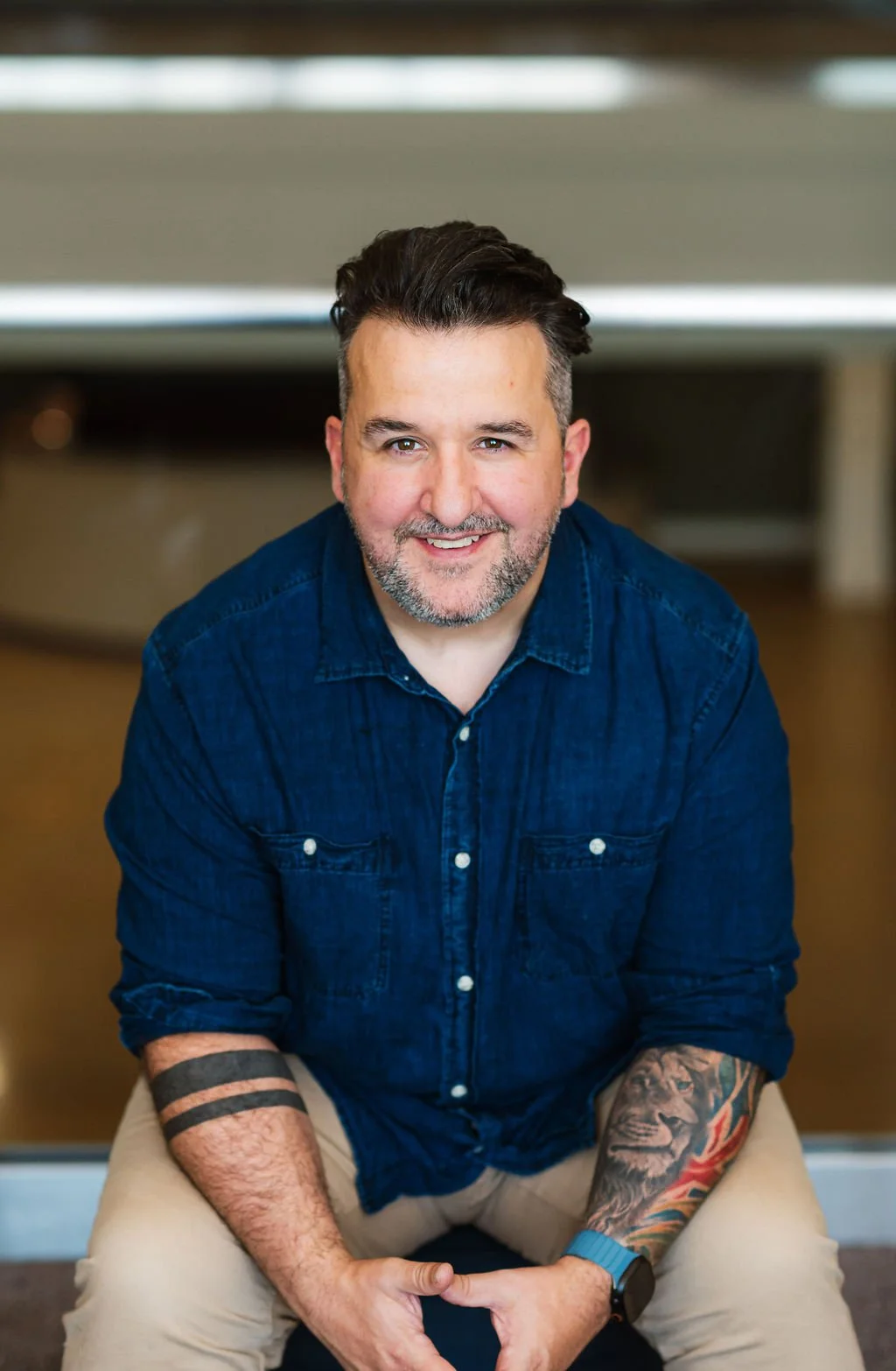 A smiling man with dark hair, beard, and tattoos on his right arm, wearing a blue denim shirt and beige pants, sitting indoors with a blurred background.