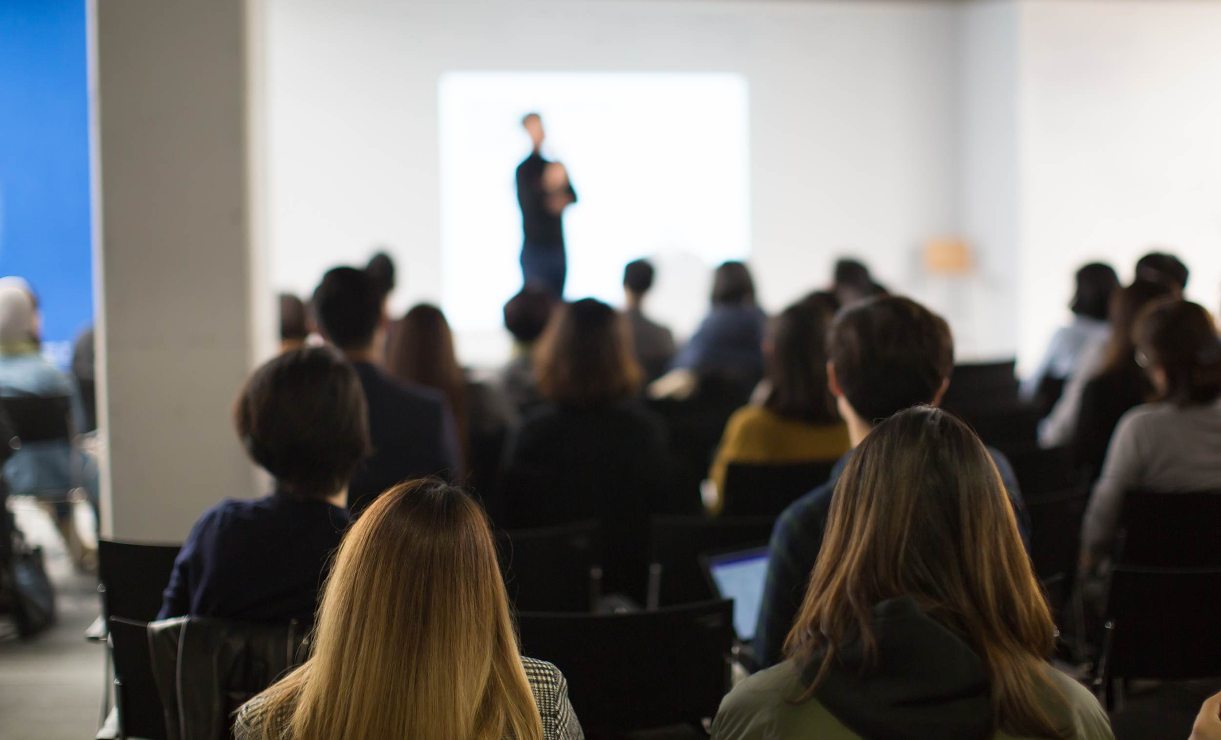 Audience attending a presentation in a conference room with a speaker at the front, blurred background.