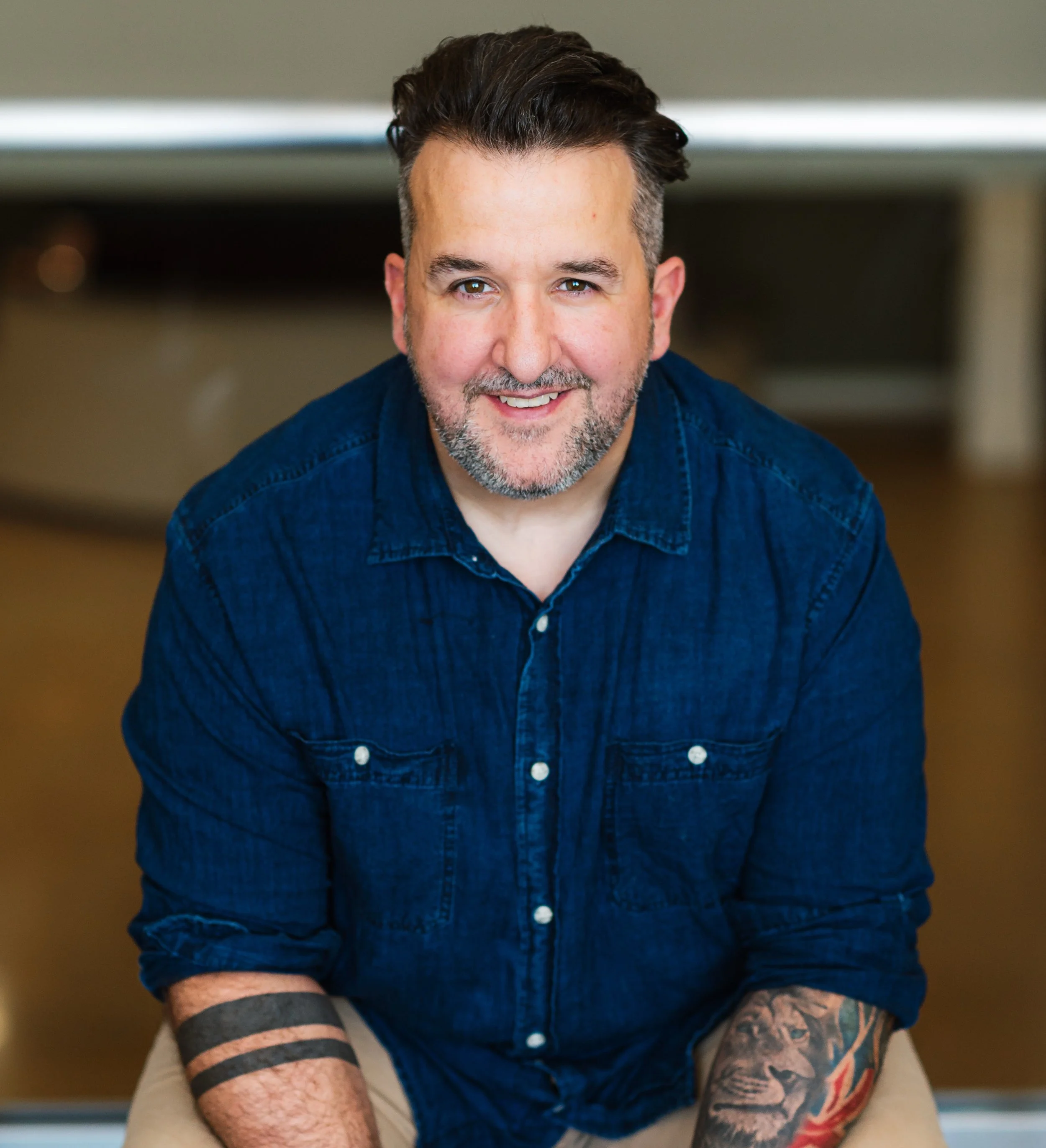 A smiling man with dark hair, a beard with gray, wearing a blue denim shirt, sitting indoors with a blurred background.