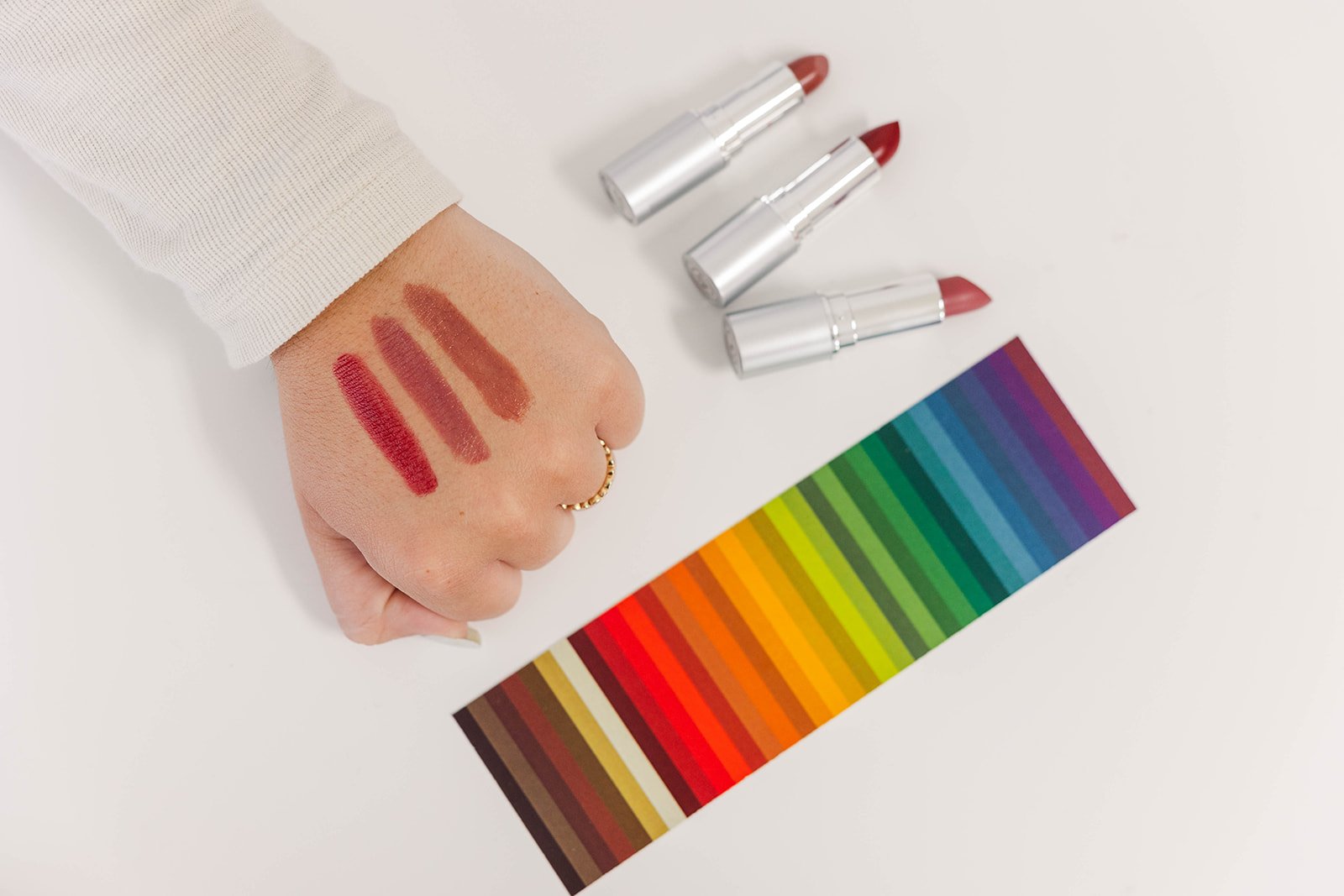 Swatches of red, pink, and brown lipstick on a person's hand, three lipstick tubes, and a color palette with rainbow colors on a white surface.