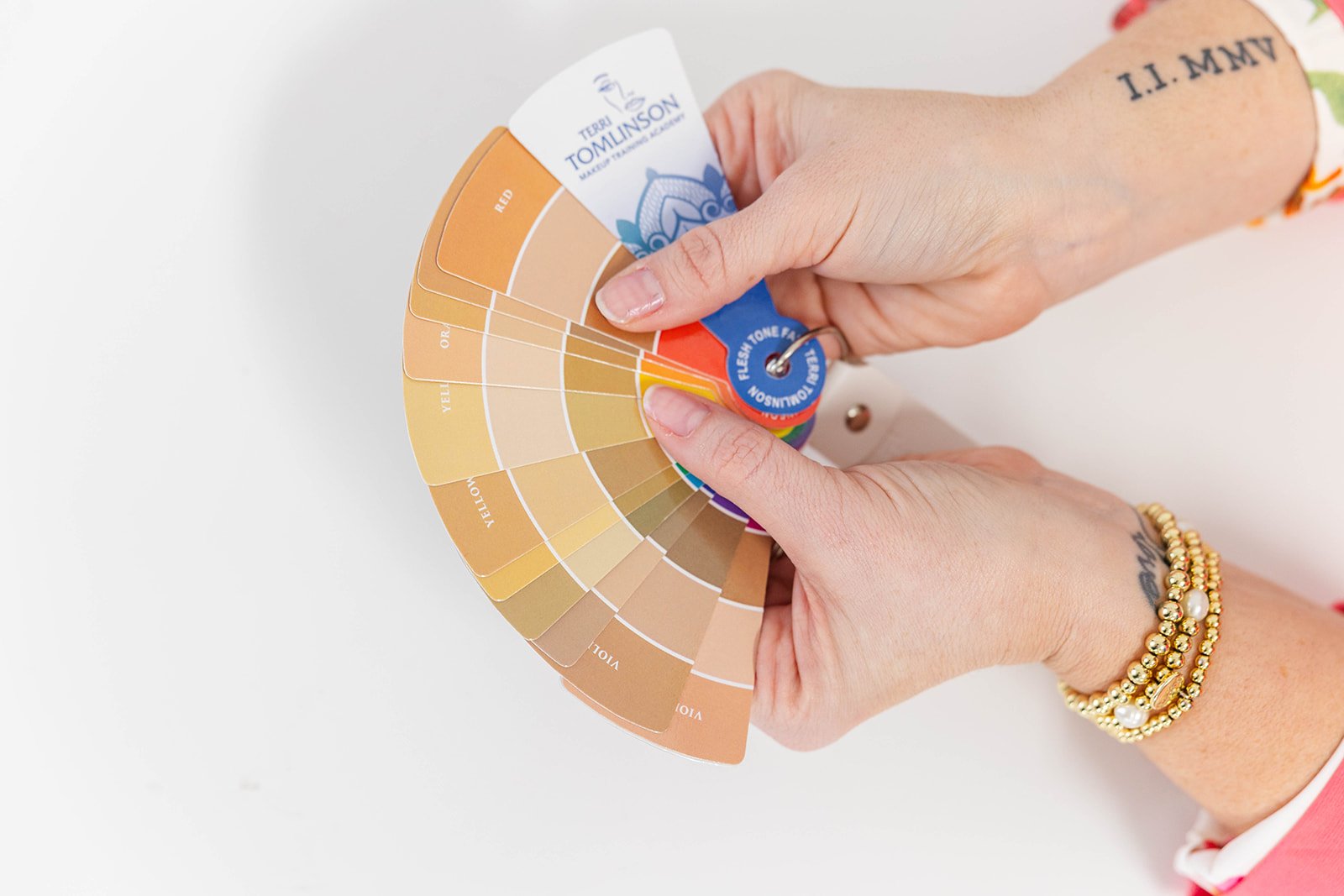Person holding a color palette fan with various shades of flesh tones, labeled with color names. The person is wearing bracelets and has a tattoo on their arm.