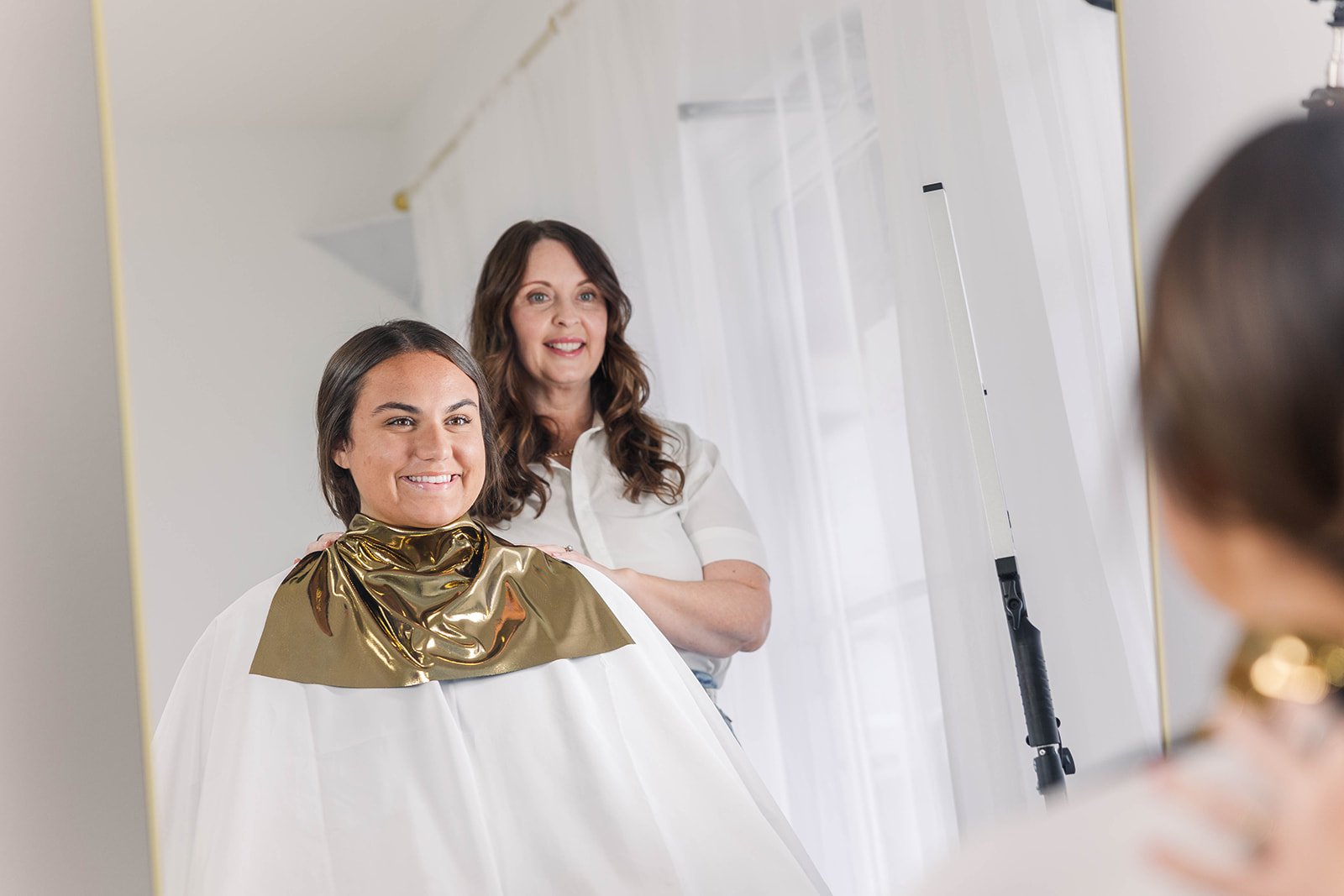 A woman getting a haircut or styling at a salon, sitting in a salon chair with a gold cape, while a stylist or friend looks on, and a mirror reflects their faces.