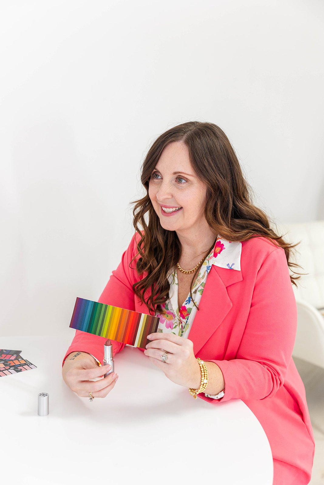 A woman with brown hair, wearing a pink blazer and floral blouse, sitting at a white table, holding a lipstick and a color palette, smiling. There are makeup palettes and a lipstick tube on the table.
