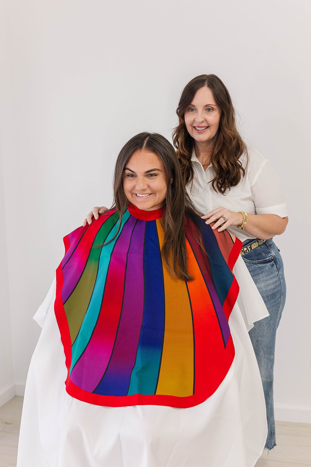 A woman getting a haircut is sitting in a salon chair with a colorful cape, while a stylist stands behind her smiling, dressed in casual white top and jeans.