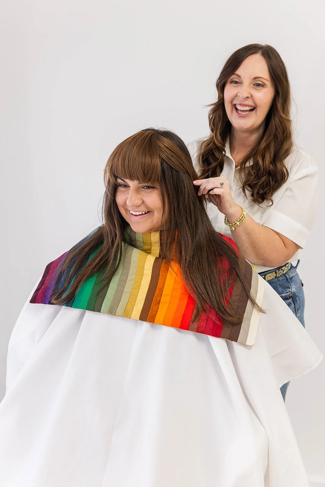 A woman with brown hair getting a haircut from a hairstylist with brown hair, both smiling in a salon.