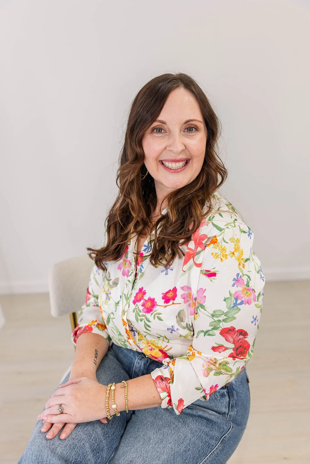 A woman with long wavy brown hair, smiling and wearing a white floral blouse and blue jeans, sitting on a white chair against a plain white background.