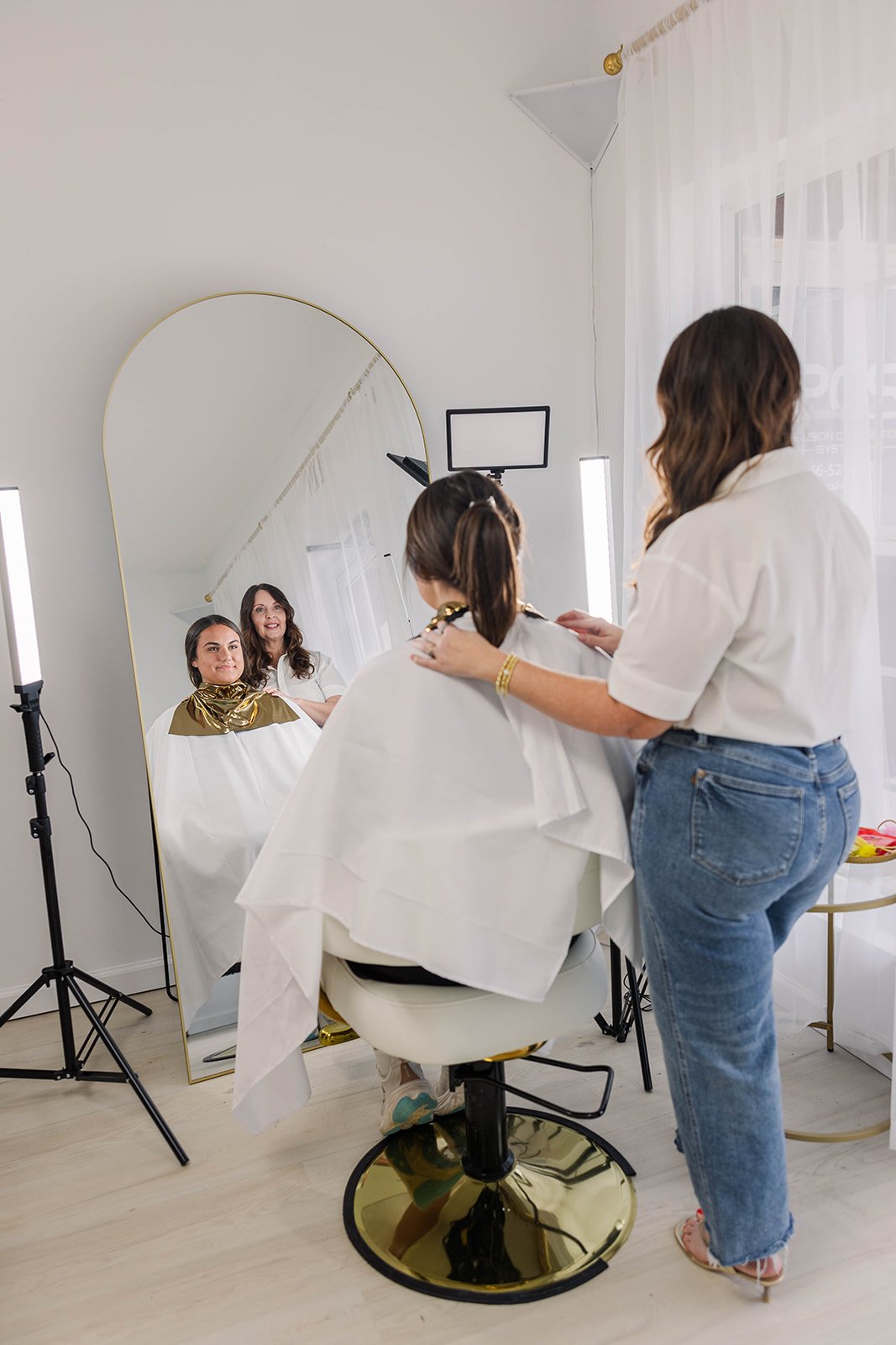 A woman getting a hairstyling or makeup service in a salon, with her reflection visible in a large mirror, while a stylist works on her, illuminated by professional lighting.