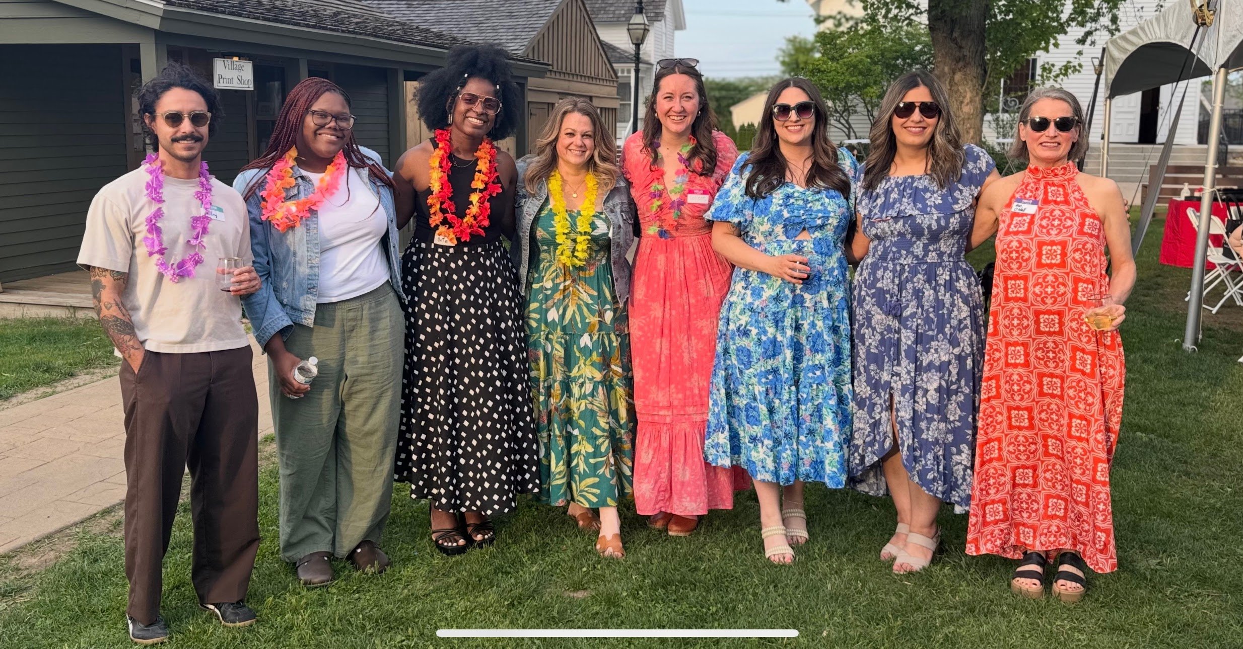 Group of nine people standing outdoors on a lawn, smiling, wearing colorful summer dresses and casual outfits, some with sunglasses and leis, in front of a building and trees at a festive gathering.