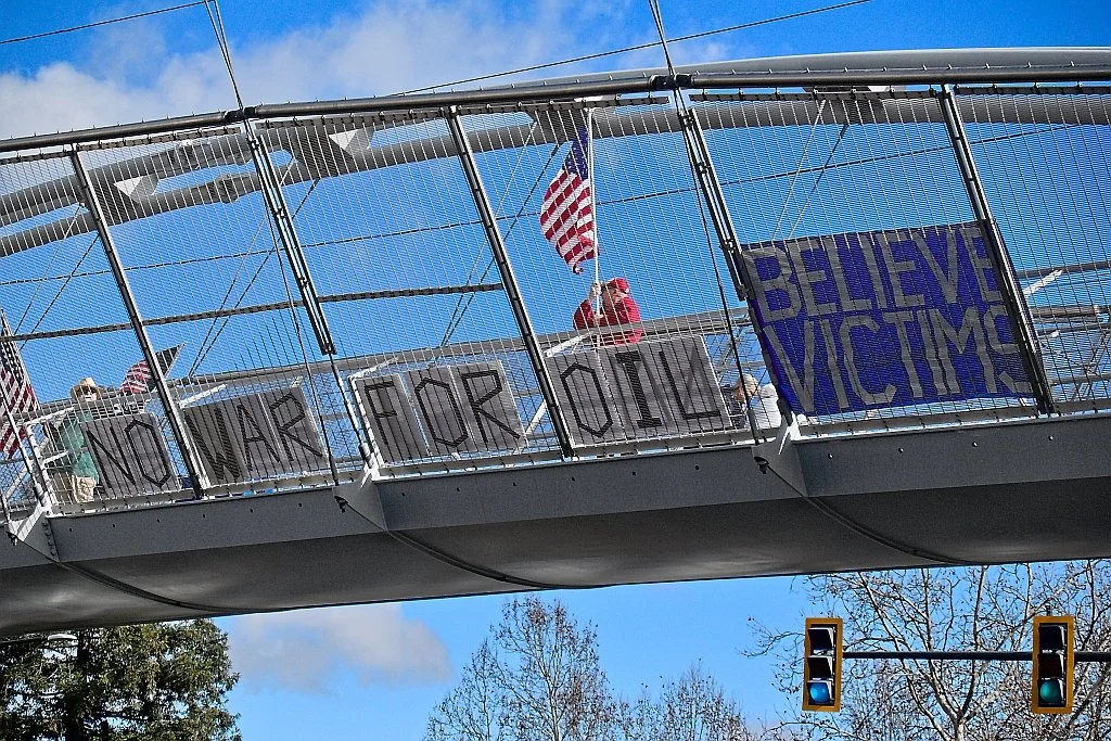 Pedestrian bridge with "No War For Oil" and "Believe Victims" signage, protester holding American flag