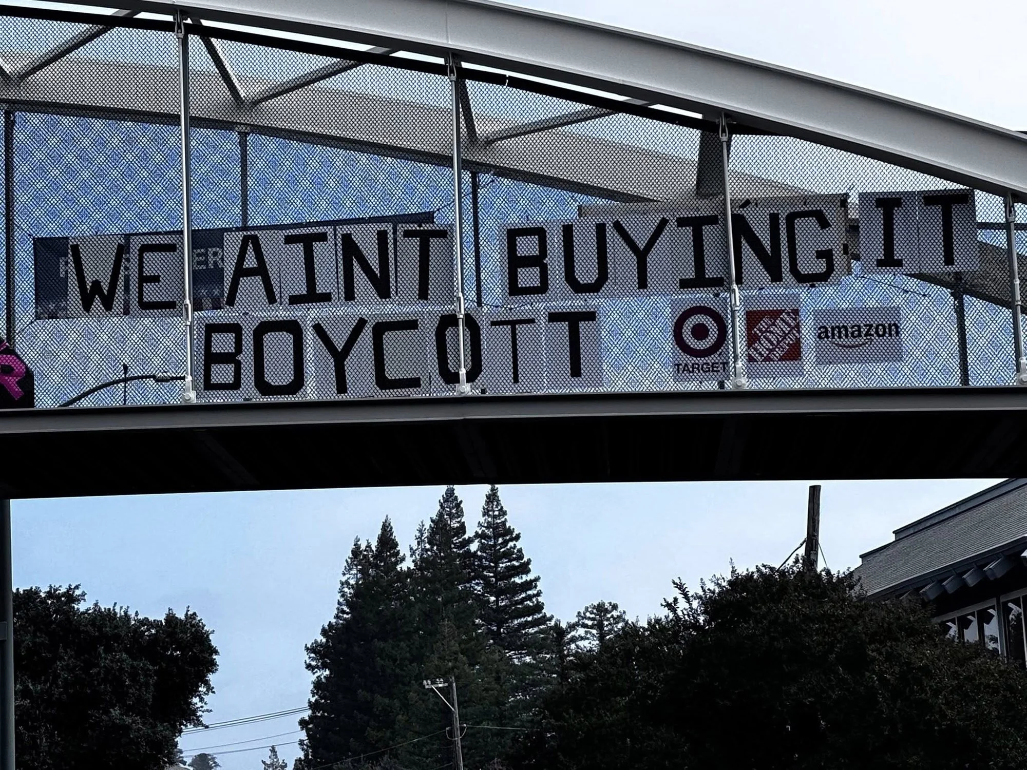 A "We Ain't Buying It Boycott" sign with Target, Home Depot and Amazon logos on a pedestrian bridge.