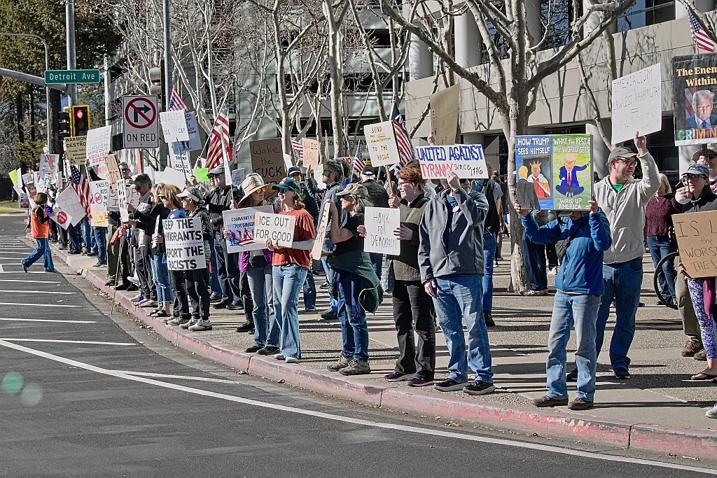 Protesters hold signs on a corner in front of a large modern building.