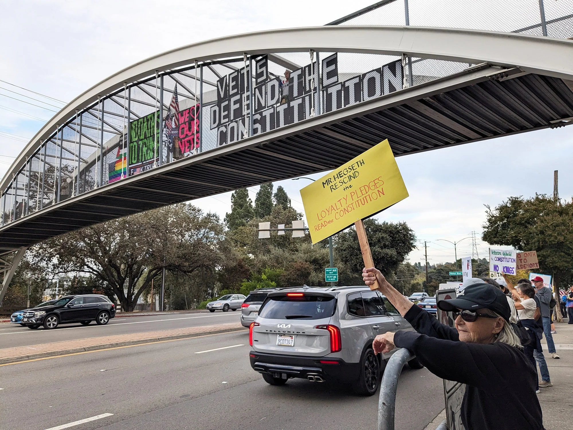 Overpass signage on YV pedestrian bridge with protesters holding signs on the sidewalk below
