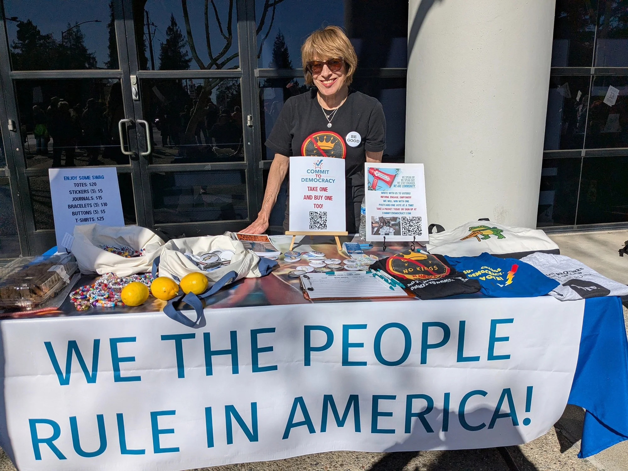A woman stands at an information table with "We the people rule in America!" sign.
