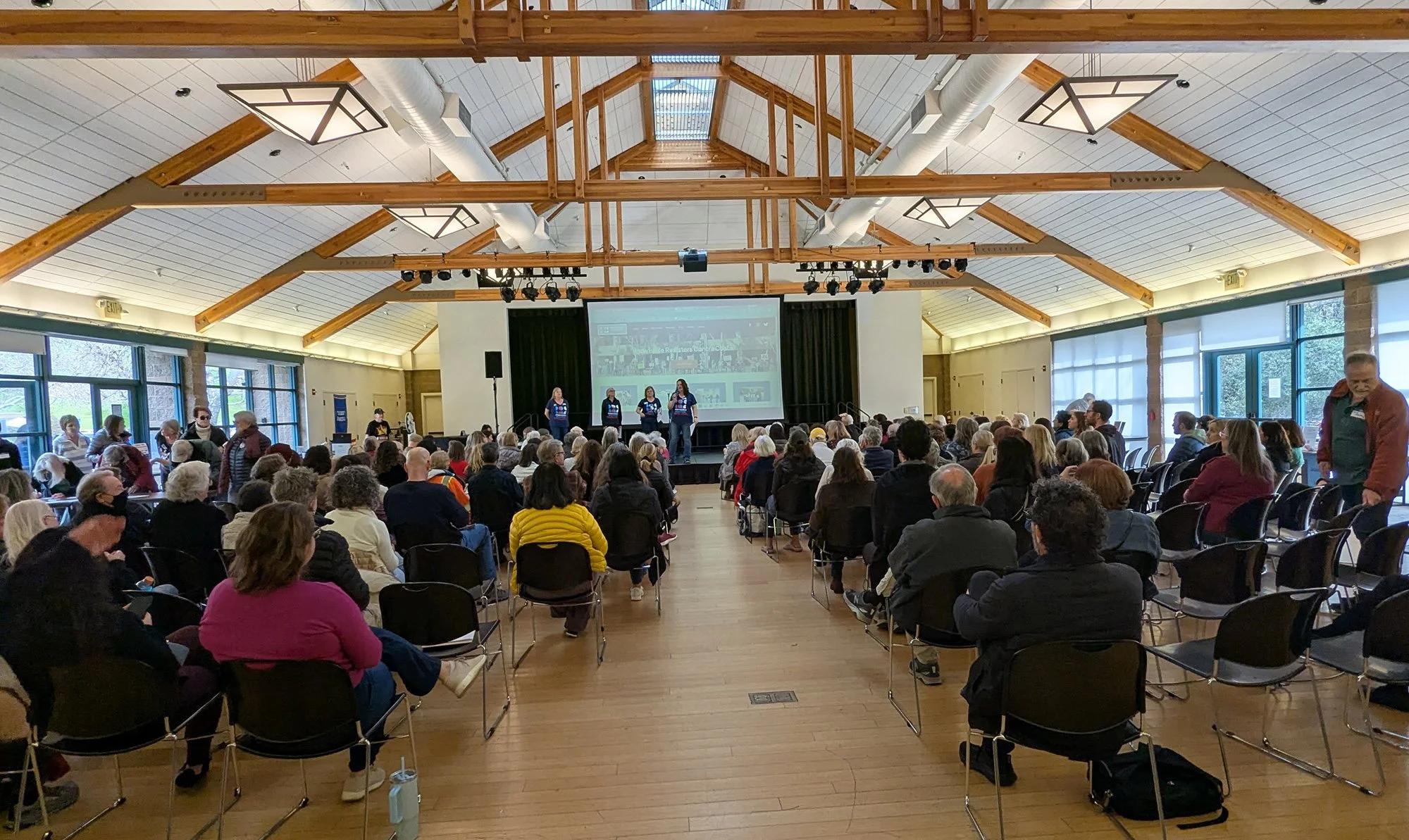 A large membership meeting seated, with speakers in front of a screen.