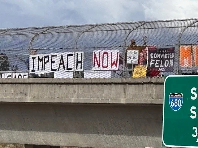 Signage on a pedestrian bridge that reads "Impeach Now" and "Convicted Felon" among others.