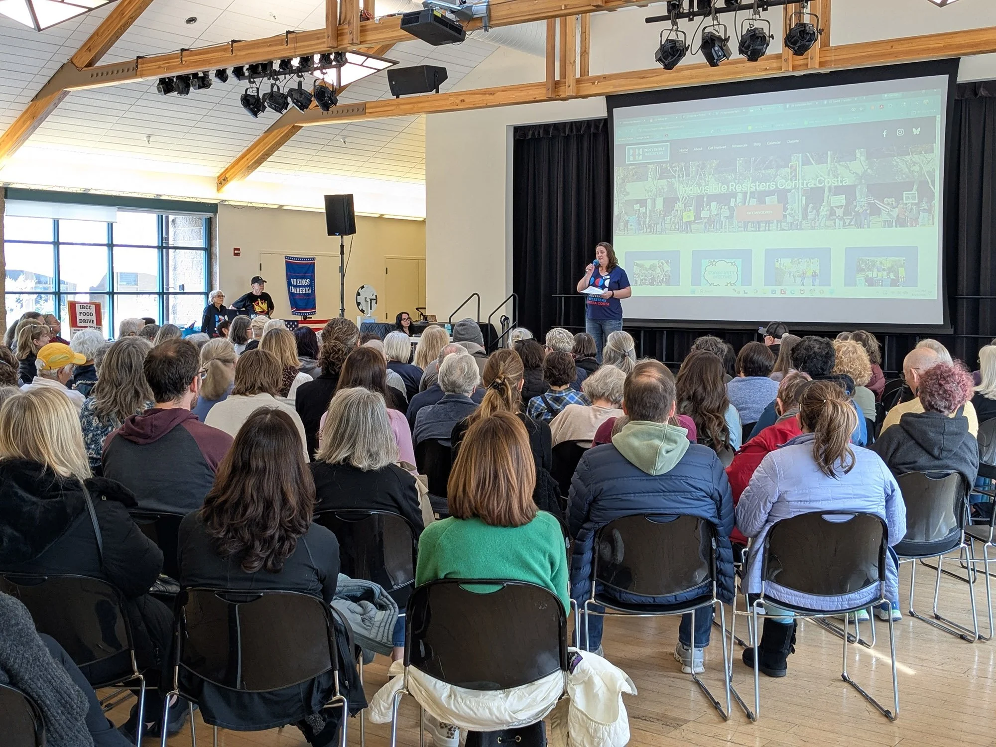 A large group of people watch a woman speak onstage at a community meeting.