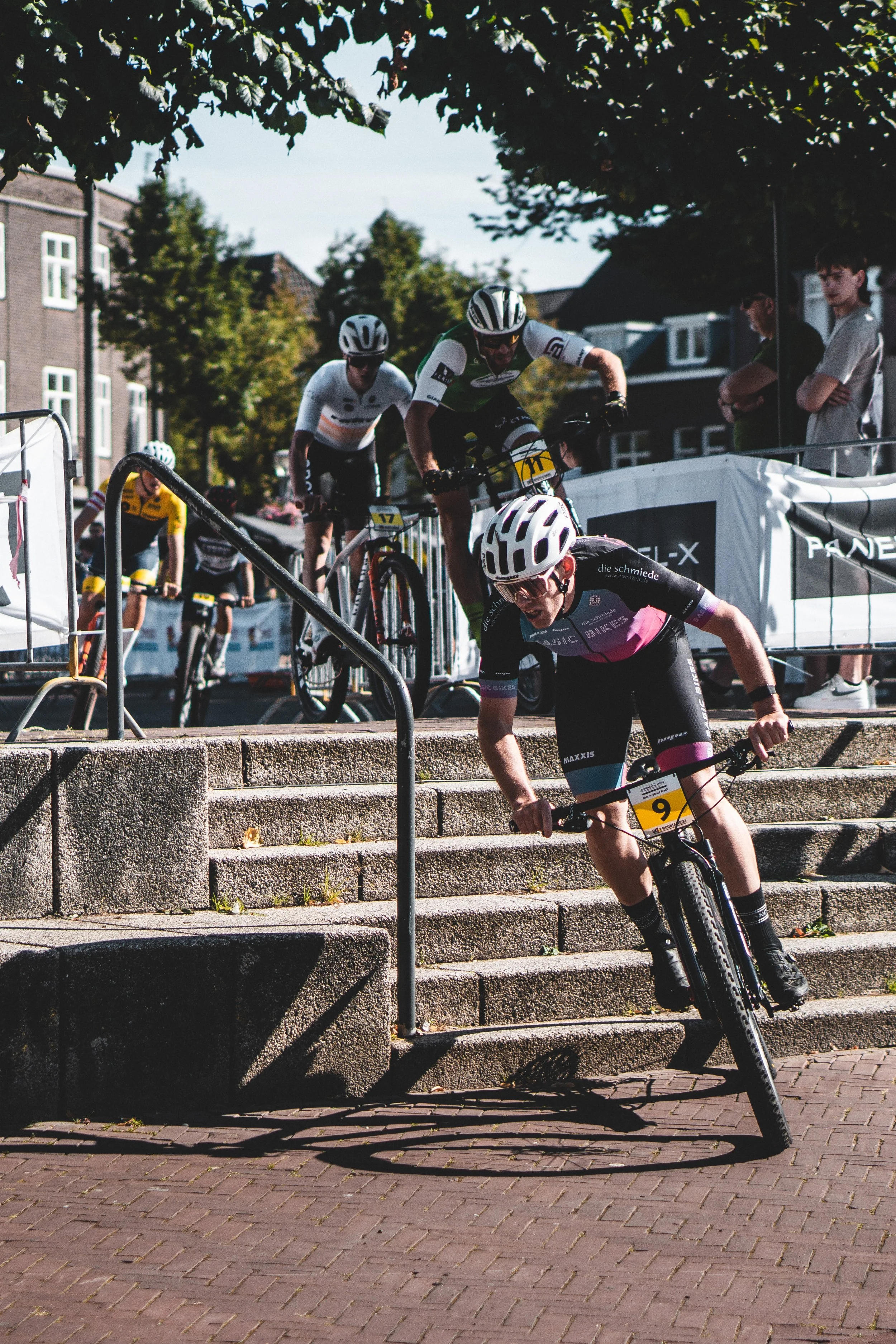 Motorradfahrer bei einem Wettkampf fährt die Treppe hinunter, während andere Radfahrer und Zuschauer im Hintergrund zu sehen sind.