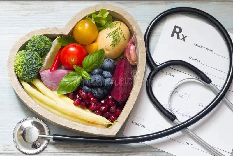 A pile of colorful fruit, with a health professionals arms writing on a clipboard, laptop is open on the table.