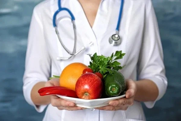 Healthcare professional holding a plate of fresh fruits and vegetables, including an apple, orange, red chili, zucchini, and cilantro.