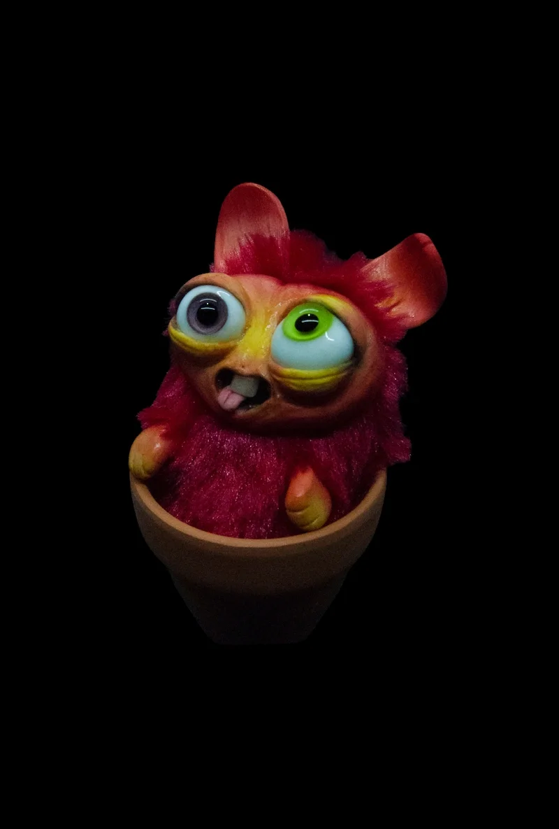 A creature with one large googly eye, one regular eye, a small tongue sticking out, and pinkish-red fur, sitting inside a small pot against a black background.