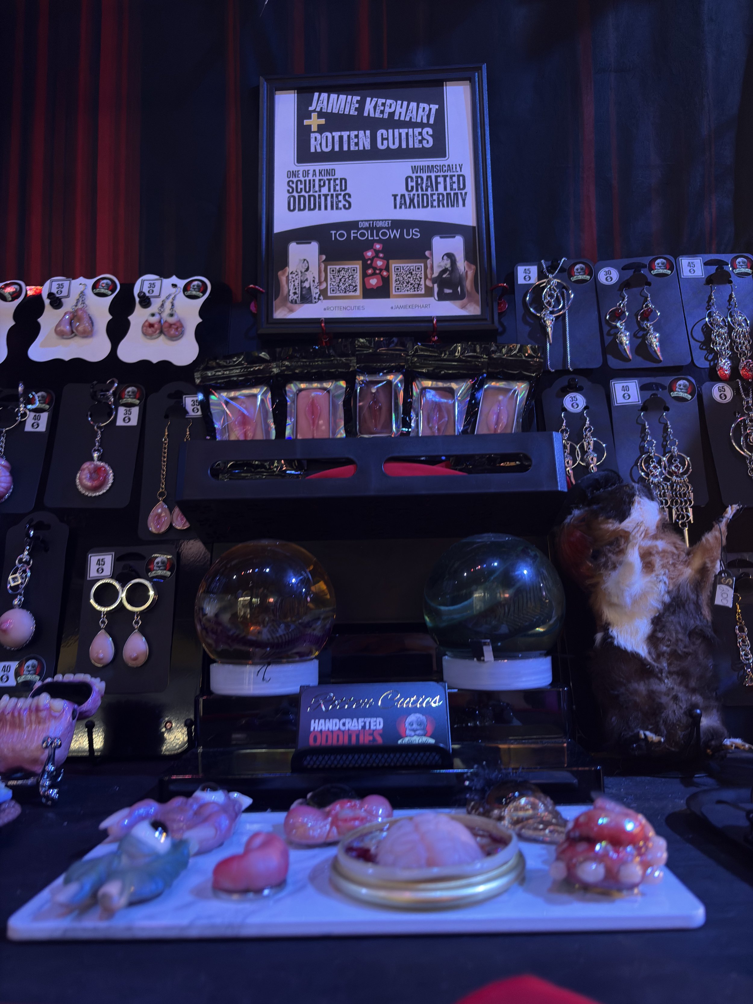 Jewelry display with earrings, rings, necklaces, and two crystal balls, featuring a sign for Jamie Kephart Rotten Cuties and handcrafted oddities