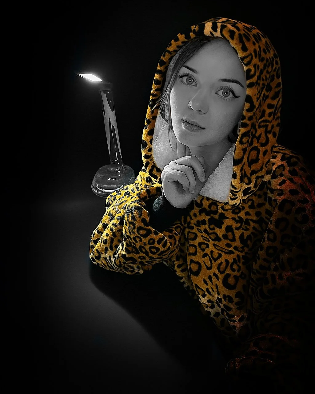 A young woman wearing a leopard print hoodie with the hood up, with a black and white filter, posing with her chin resting on her hand while looking at the camera. There is a lit candle in the background.