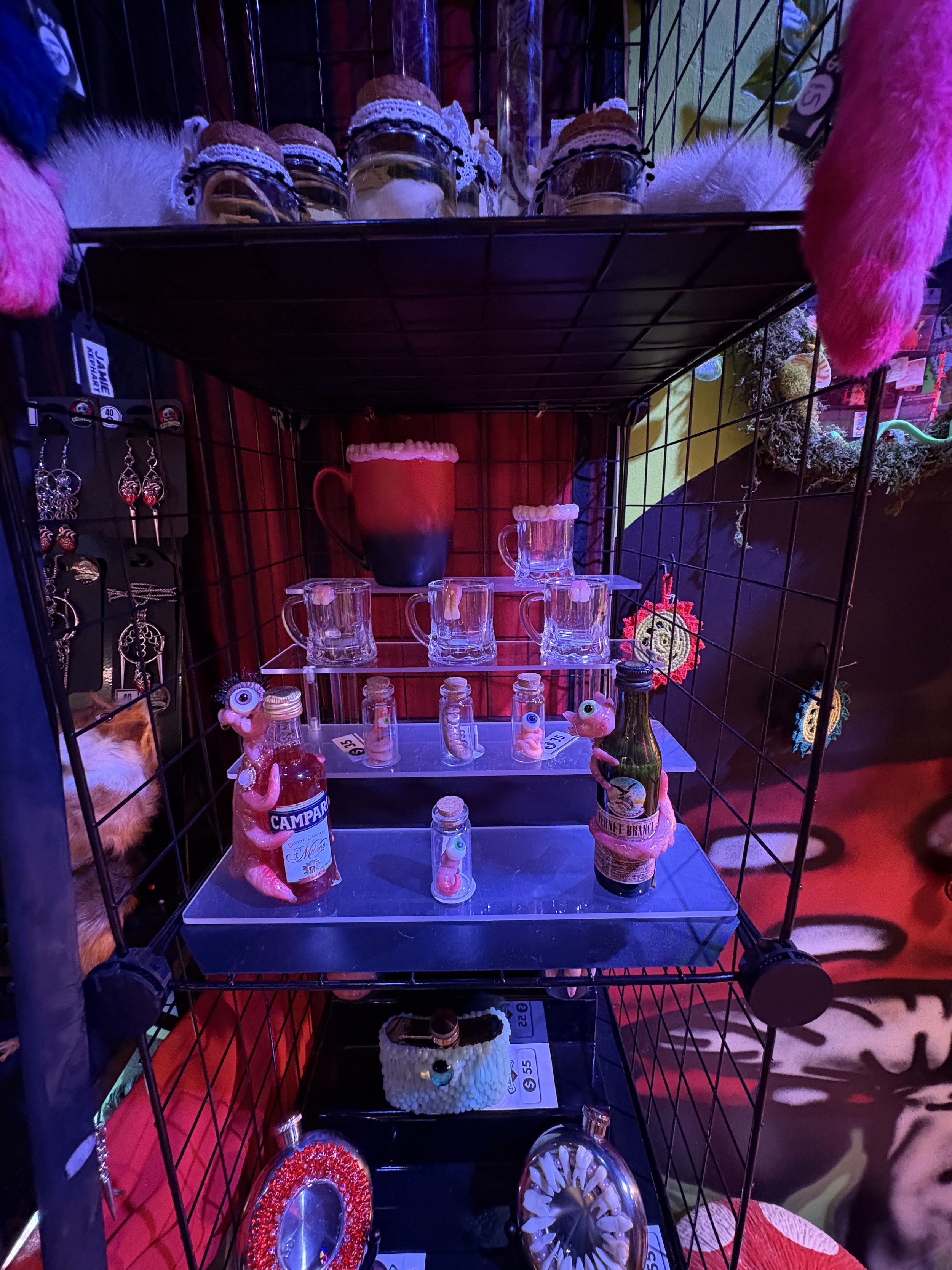 A display of holiday-themed small decorative items on a black metal shelf, including jars, glass cups with fake whipped cream, tiny bottles with quirky labels, and small plush toys, with a red and black background.