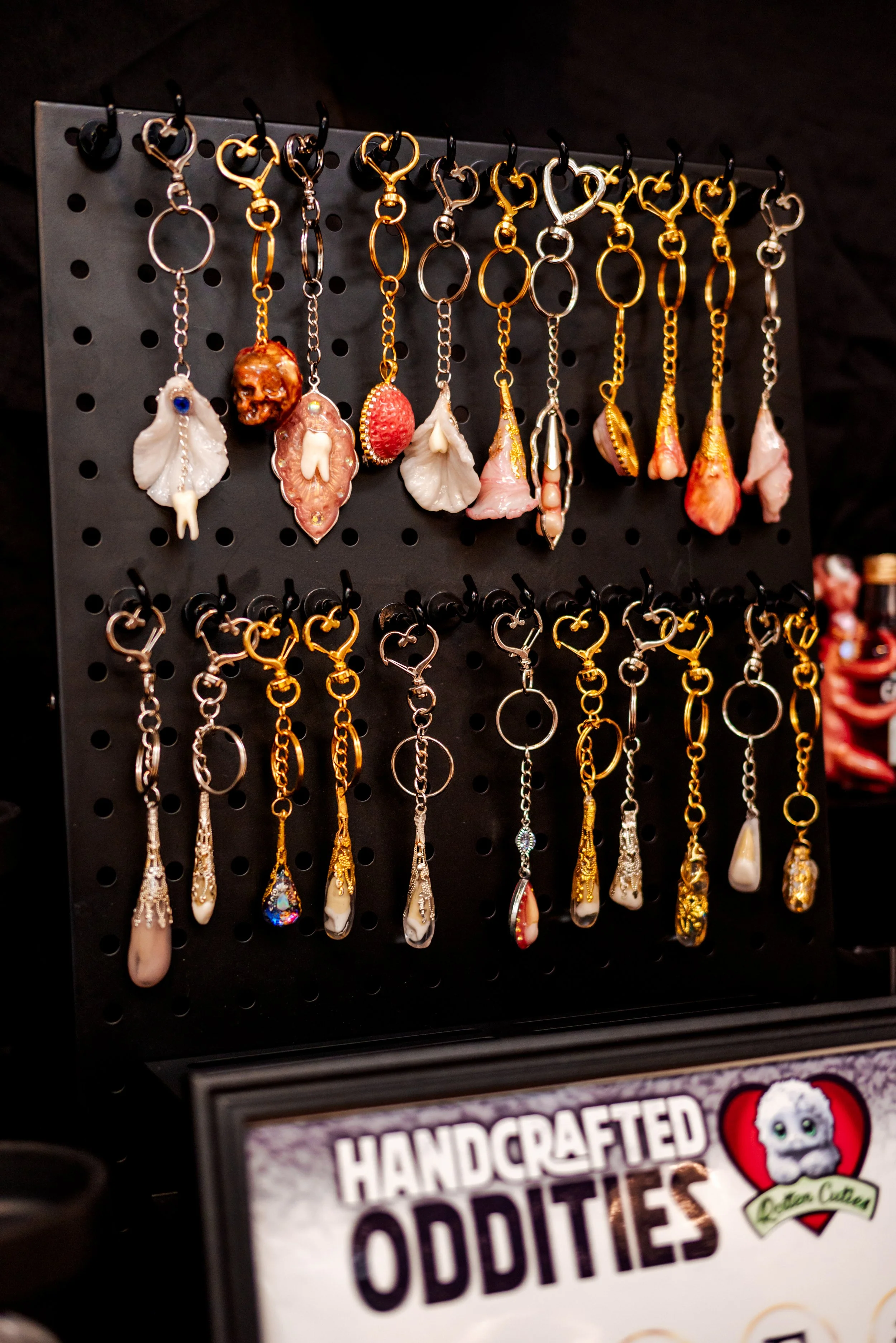 A display of handcrafted oddities, including keychains with various miniature objects like teeth, shells, and small figurines, hanging on a black pegboard.