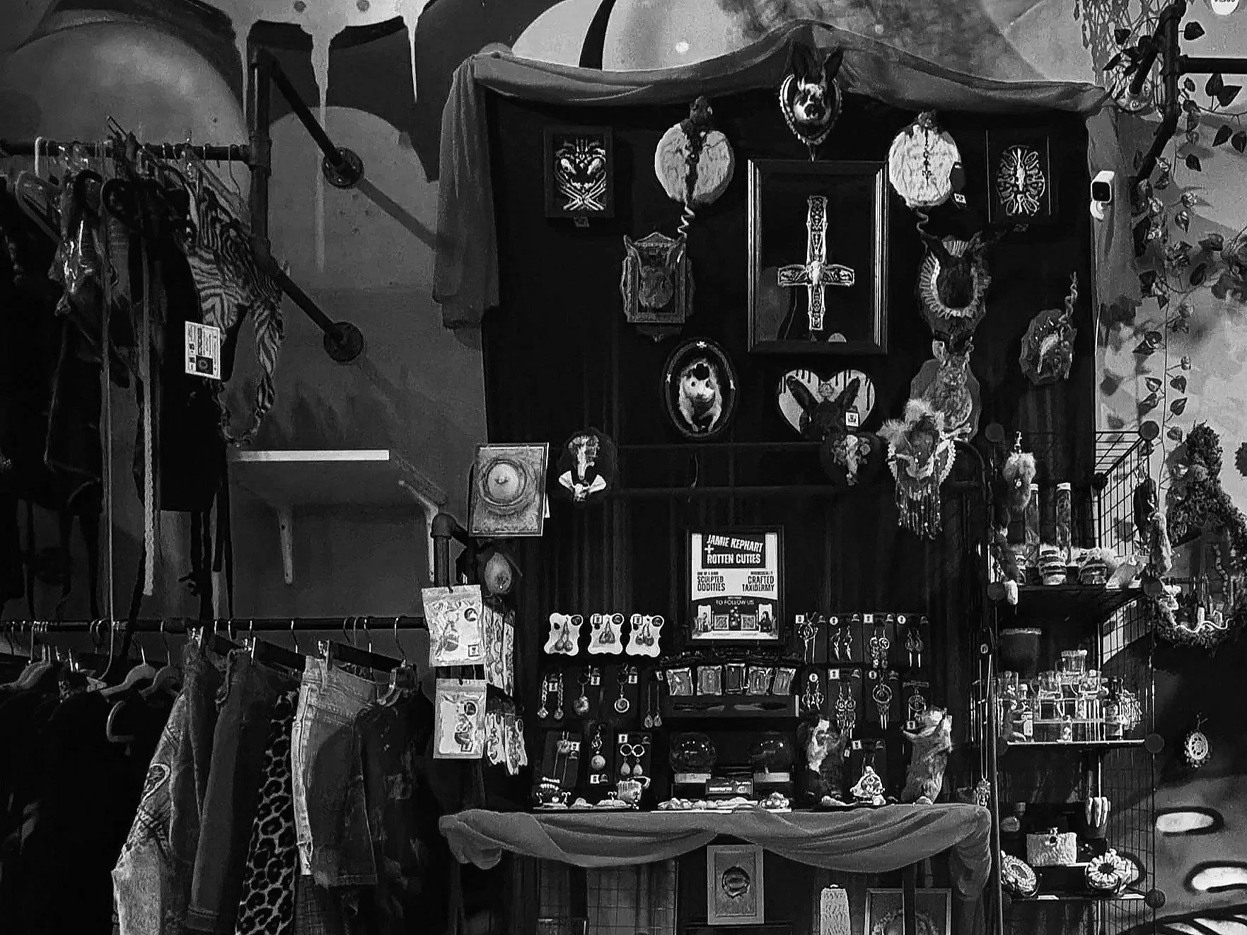 A black-and-white photo of a display shelf with various religious and decorative items, including crosses, masks, and figurines, with clothing hanging on a rack to the left.