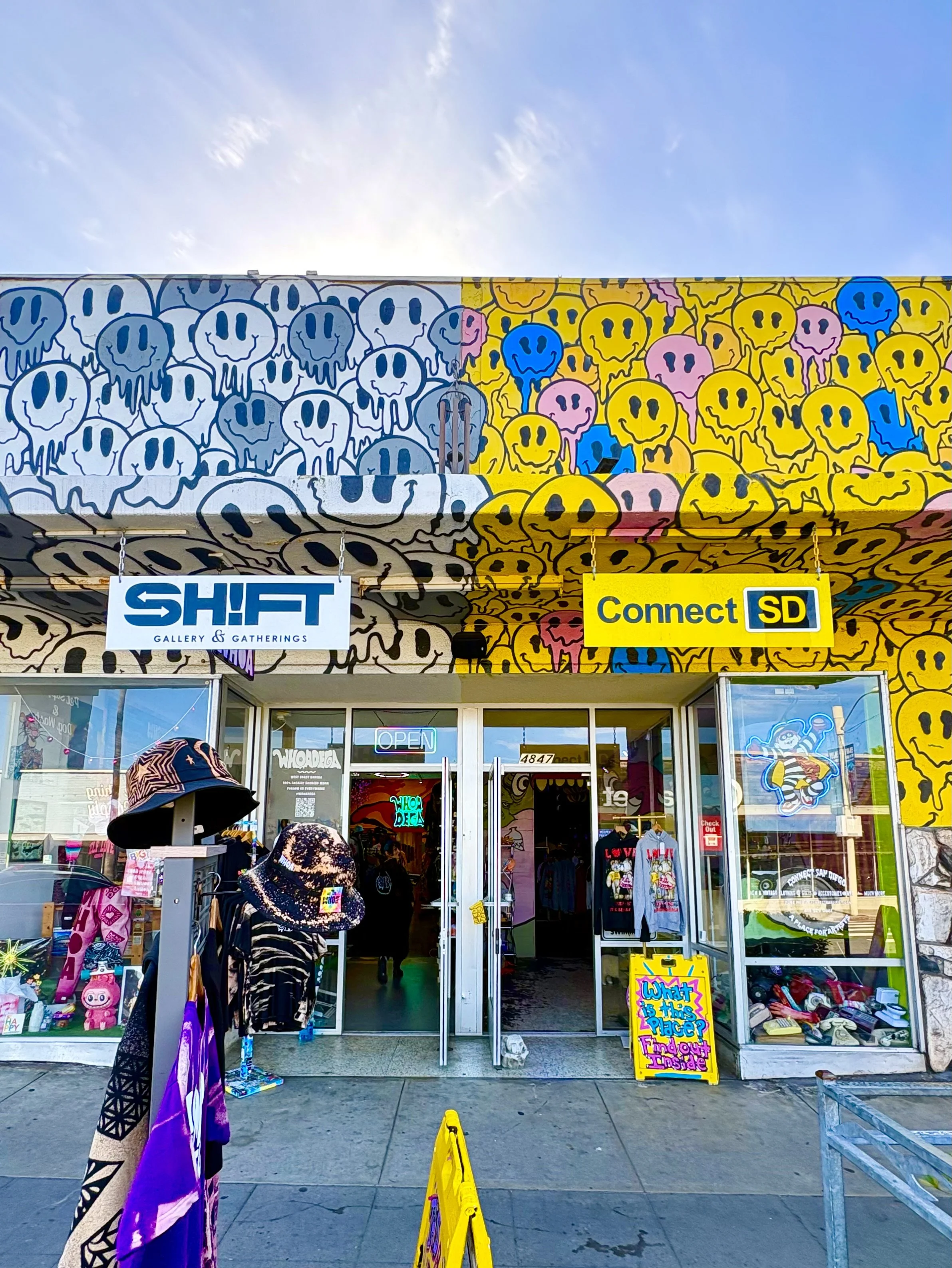 Colorful mural of smiling skulls in black, white, yellow, pink, and blue covers the top of a gallery storefront with signs that read "SHIFT Gallery & Gatherings" and "Connect SD". The storefront has glass doors and windows displaying merchandise, with outdoor decorations including hats and a signboard.