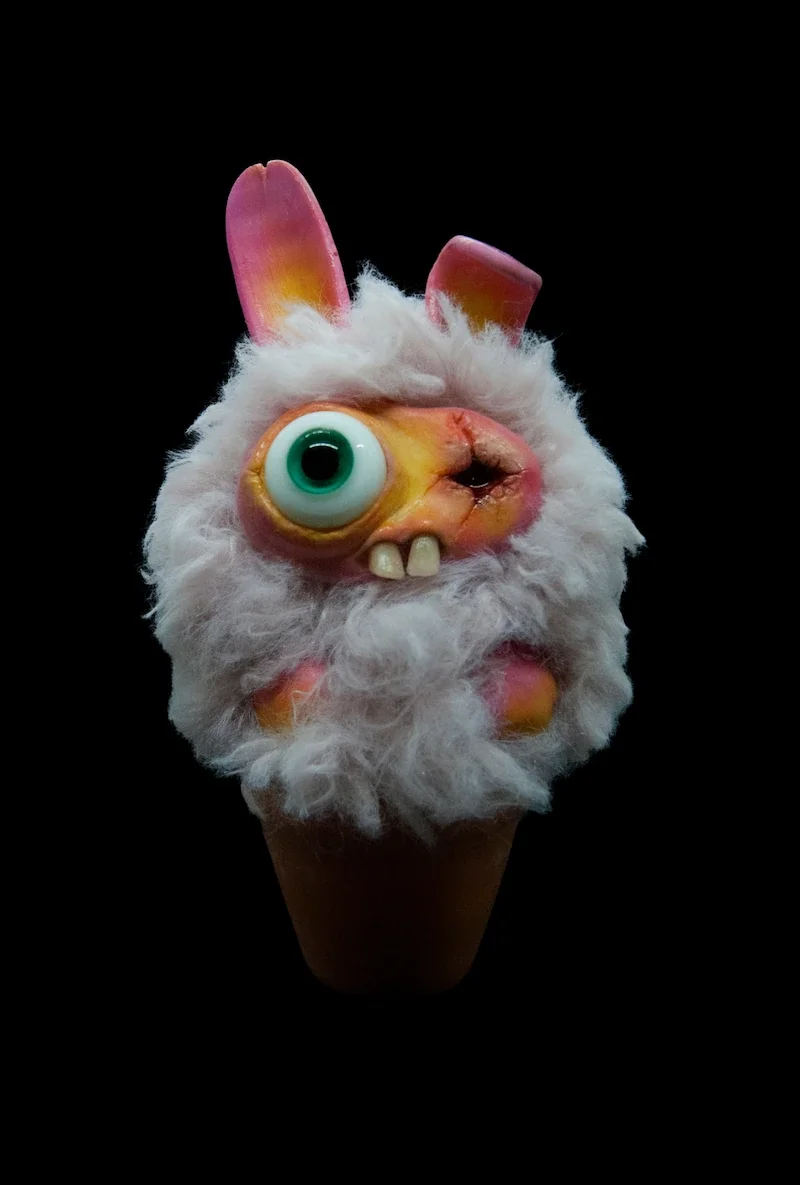 A fuzzy, surreal ice cream cone sculpture resembling a cartoonish monster with one large eye, two front teeth, pink bunny ears, and a fluffy white beard, set against a black background.