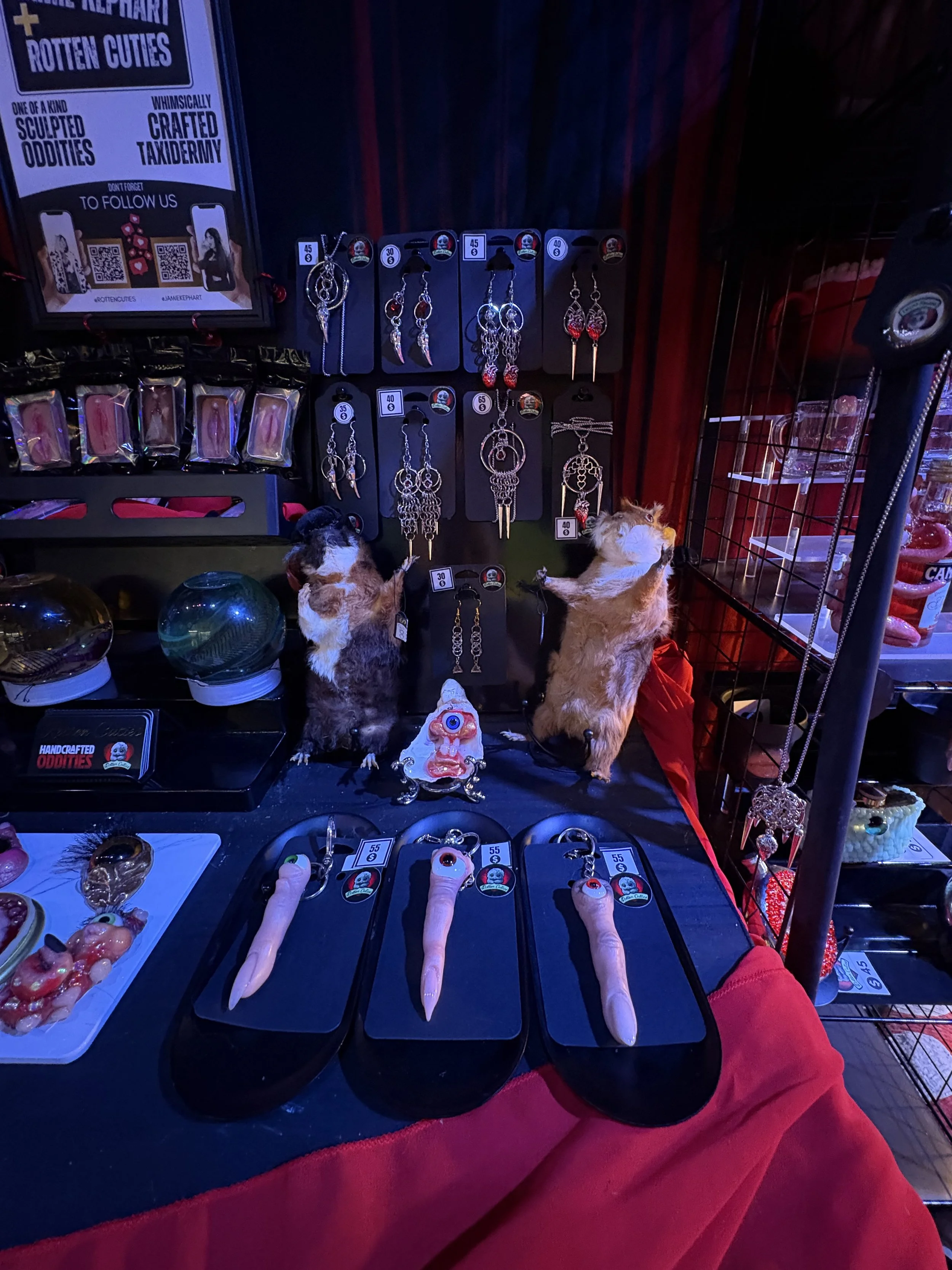 Display of Halloween-themed accessories and decorations at a booth, including spider-shaped jewelry, plush rat and mouse figures, and novelty keychains or ornaments with tentacle designs and a creepy eye, decorated with red and black colors for Halloween.